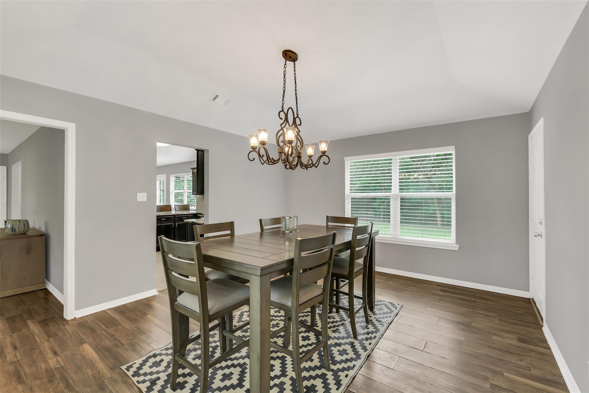 Dining room with a rectangular table, geometric rug, chandelier, and large window with blinds.