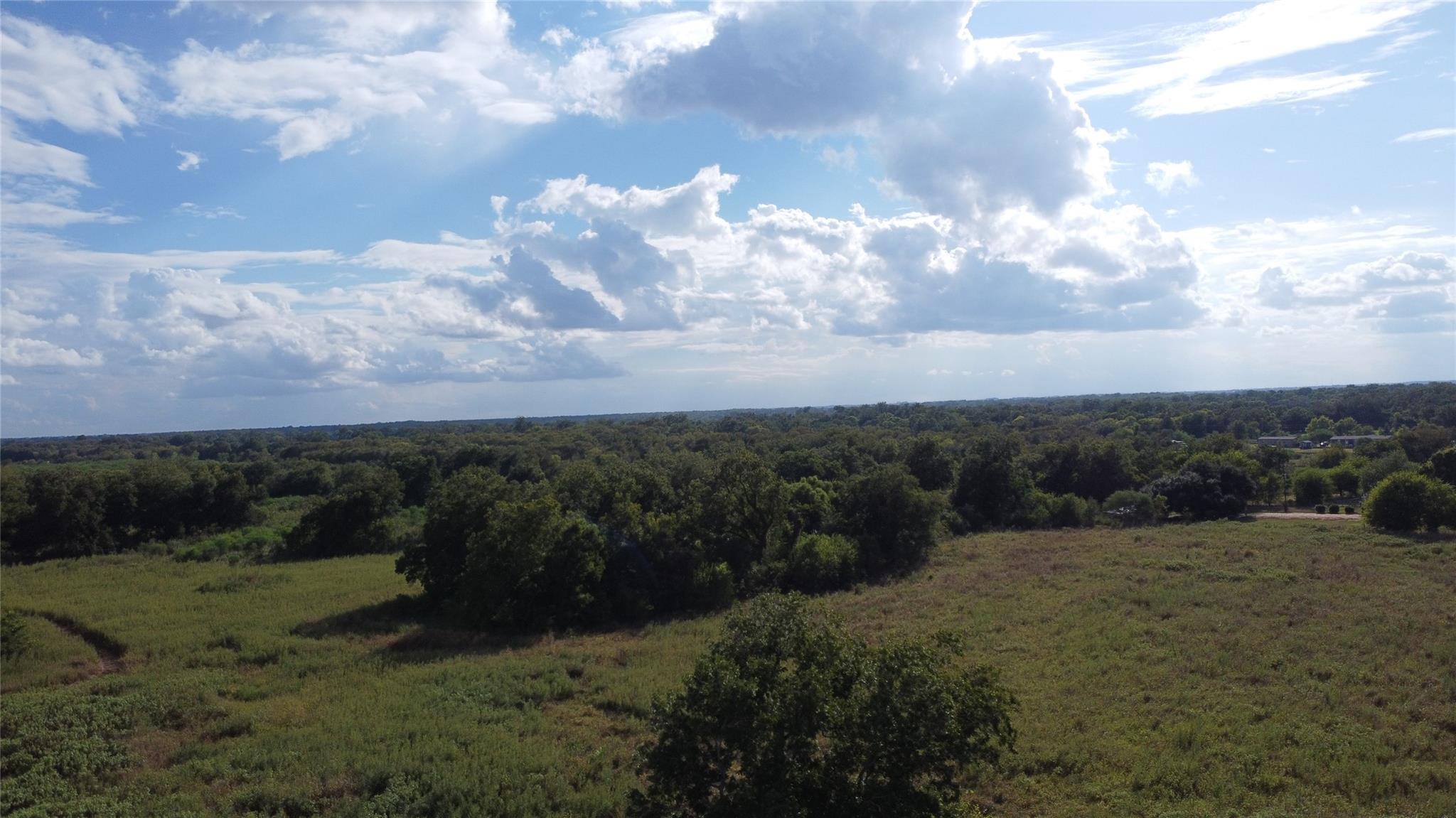 Aerial view of grassy rural land, dense trees, and distant buildings beneath a cloud-filled sky.
