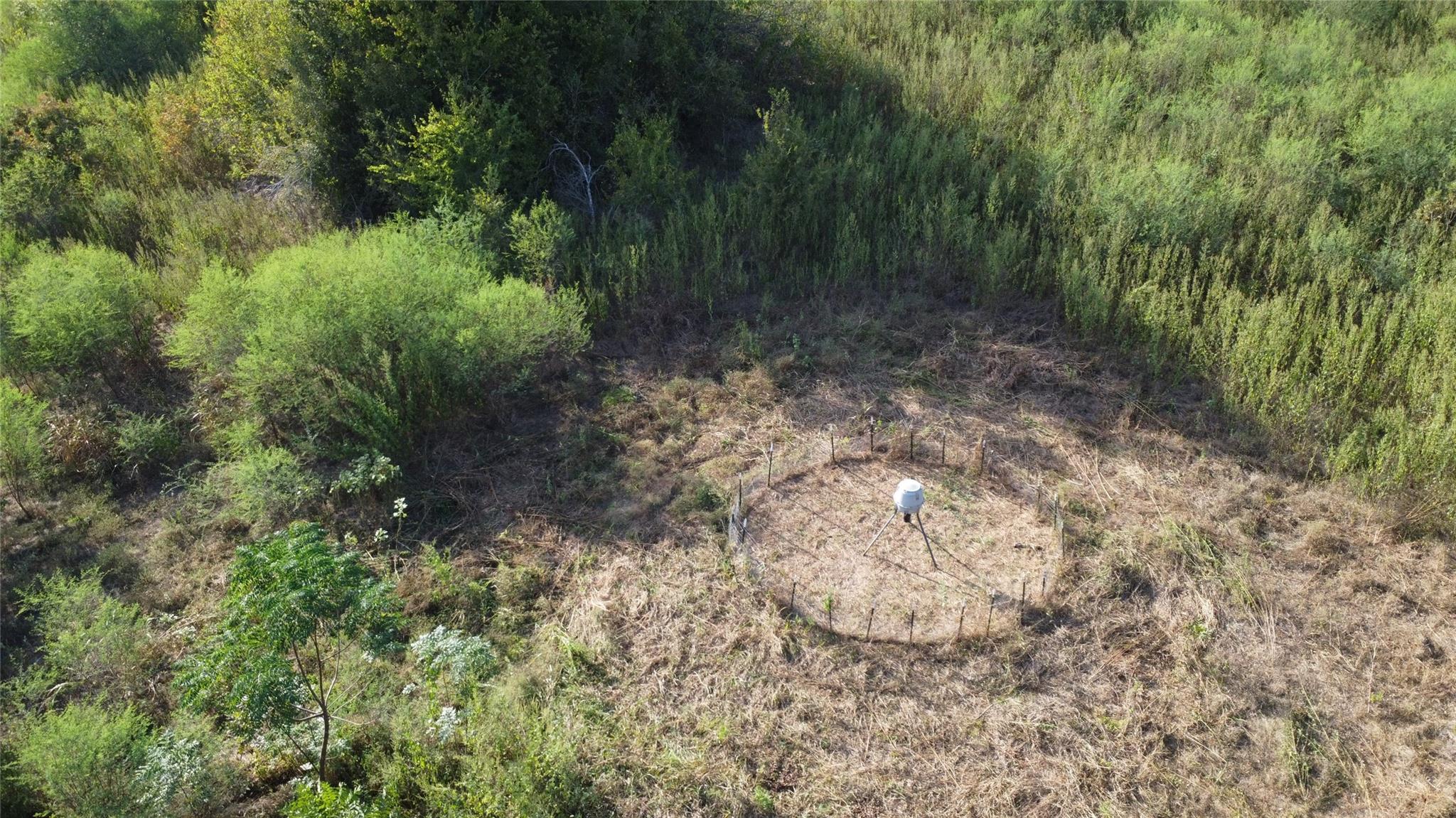 Aerial view of a brushy rural lot with a fenced clearing and a small white device.