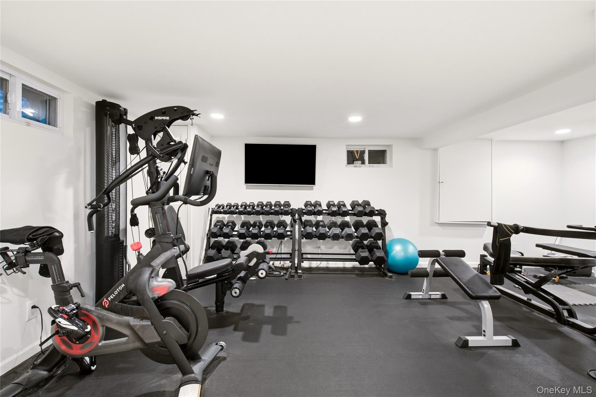 Home gym with Peloton bike, dumbbell racks, strength machine, benches, and blue exercise ball.