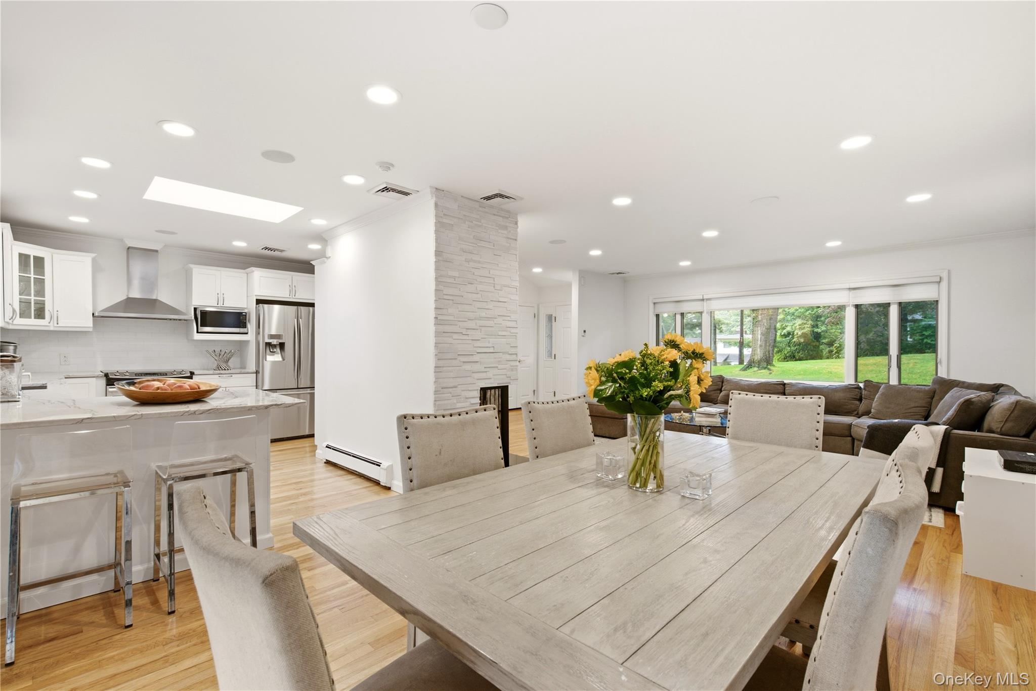 Open-plan kitchen and dining area with a large wood table, stone fireplace, and windows overlooking the yard.