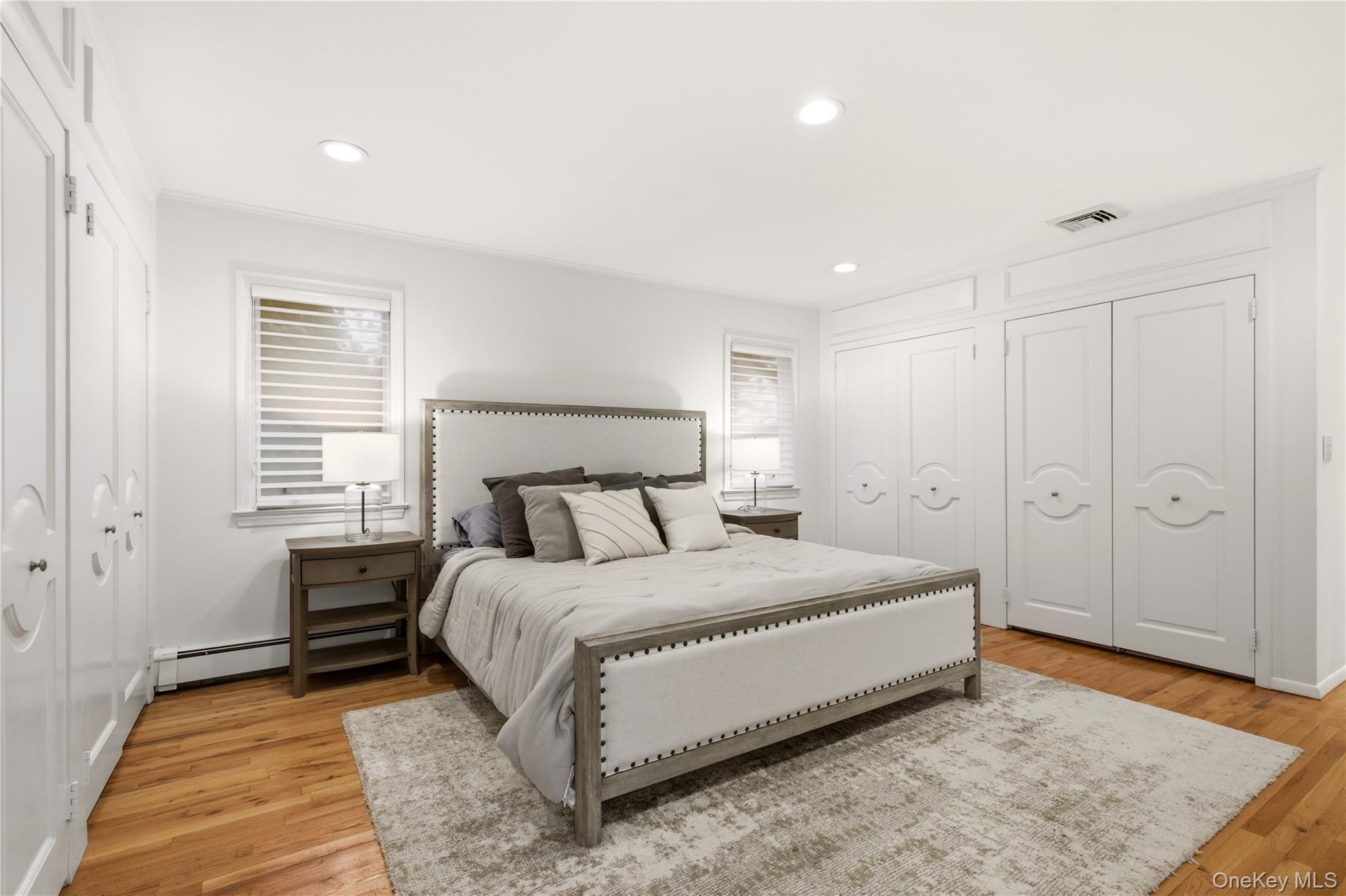 Furnished bedroom with a large upholstered bed, white paneled closets, hardwood floors, and striped window shades.