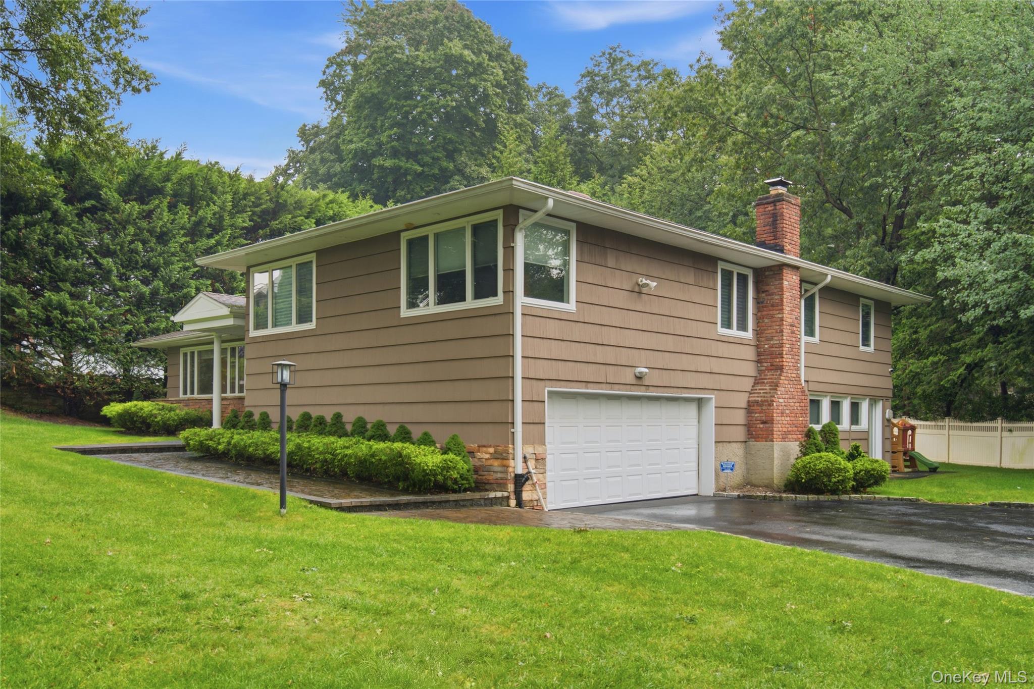 Brown split-level home with a brick chimney, attached garage, landscaped lawn, and mature trees.