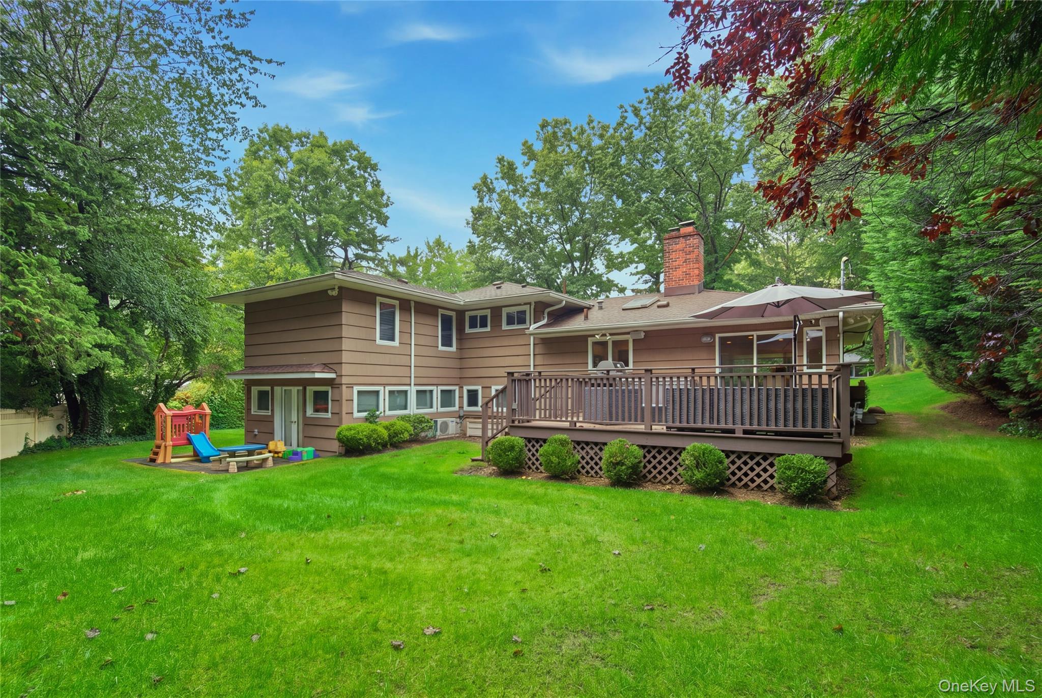 Rear of a brown-sided house with a raised deck, patio umbrella, and children's playset on a large lawn.