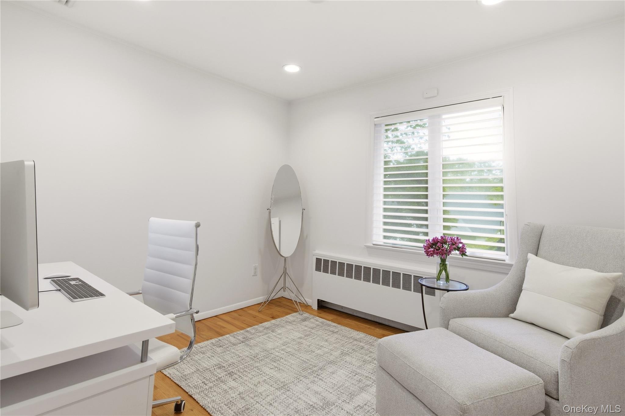 Home office with a white desk, gray lounge chair, ottoman, and window with blinds.