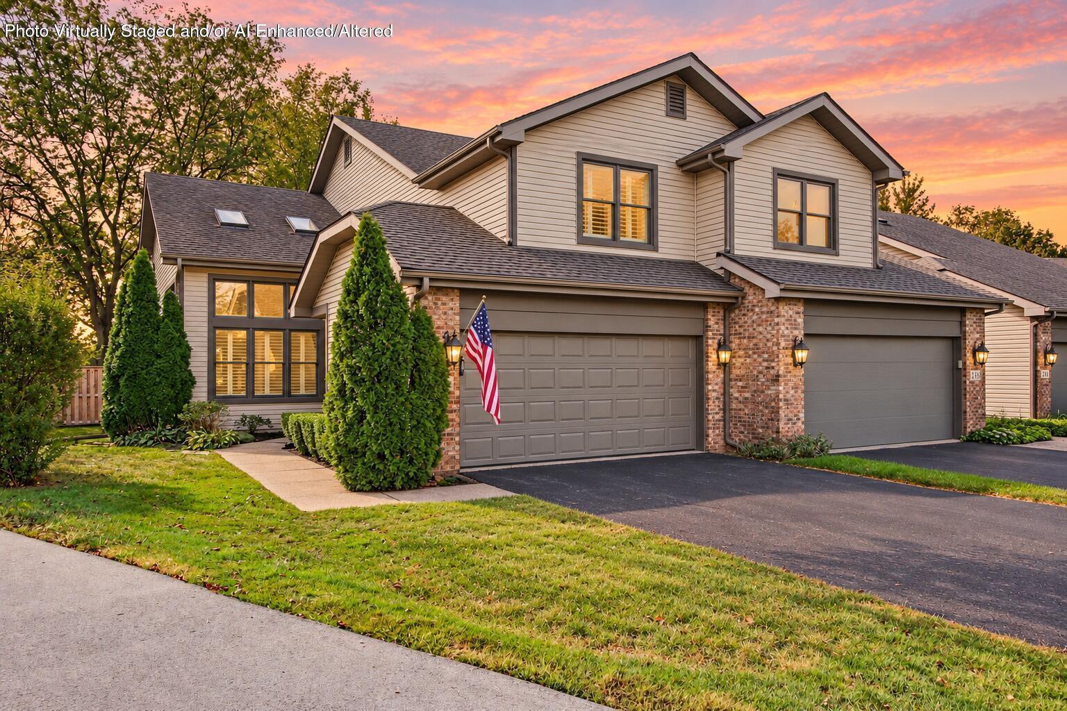 Two-story beige and brick house with a double garage, landscaped yard, and American flag at sunset.