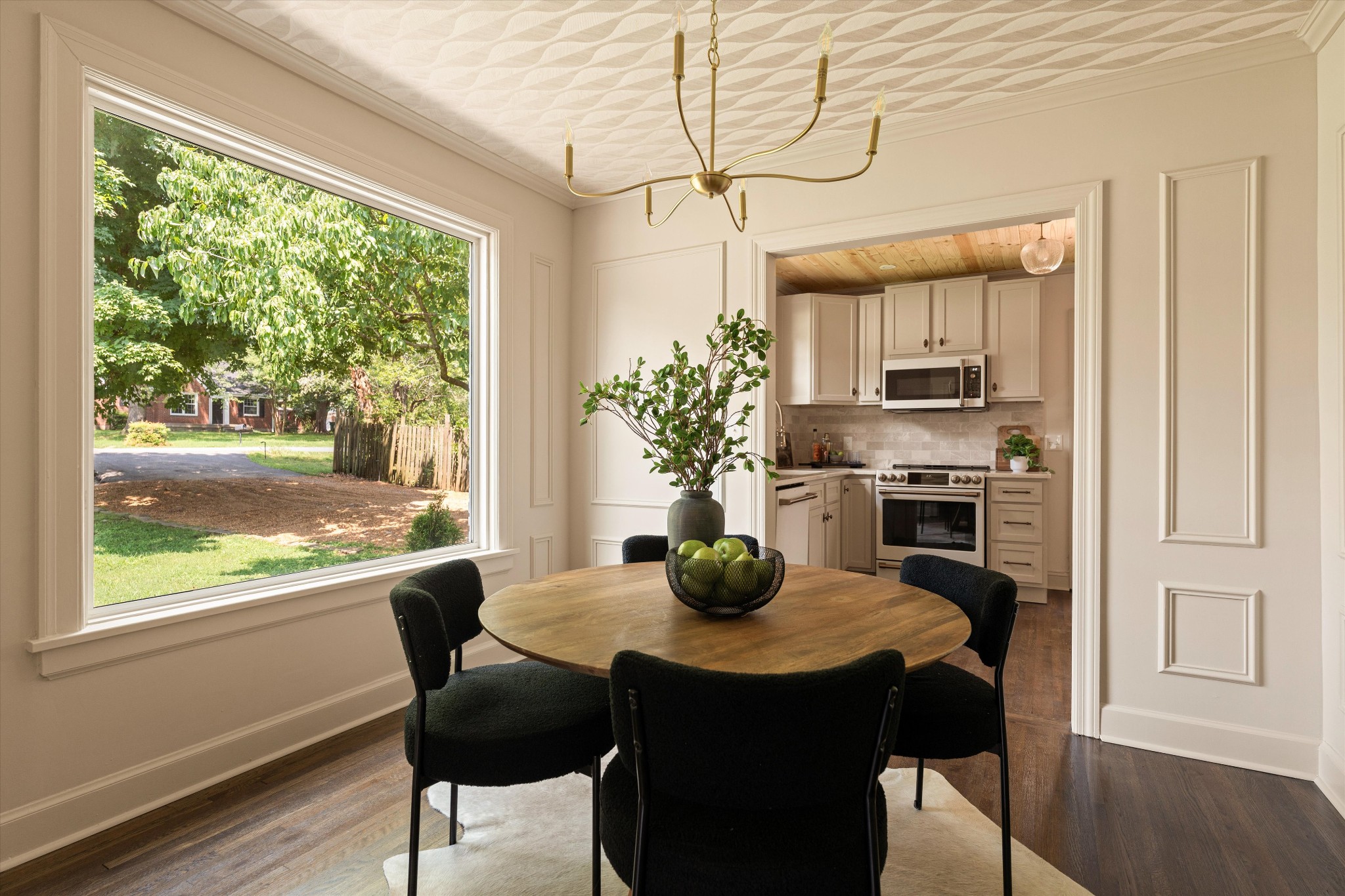 Dining room with a round wooden table, large yard-facing window, and adjoining kitchen.