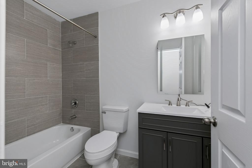 Bathroom with gray tiled tub surround, toilet, and dark vanity with mirrored cabinet.