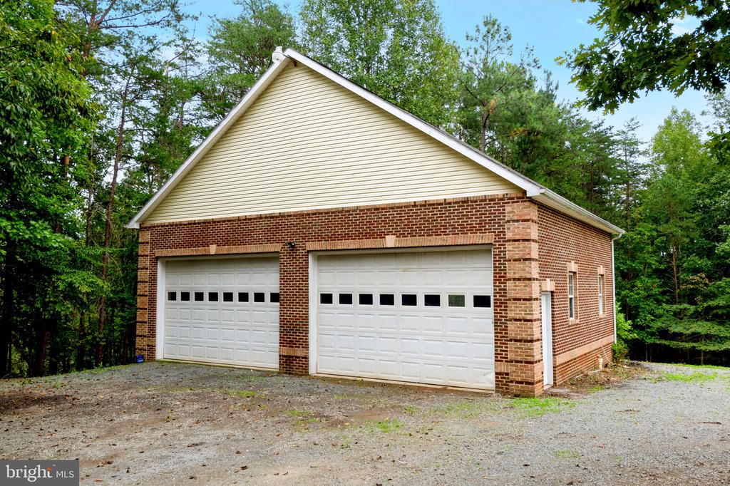 Detached brick garage with two white overhead doors beside a gravel driveway and wooded surroundings.