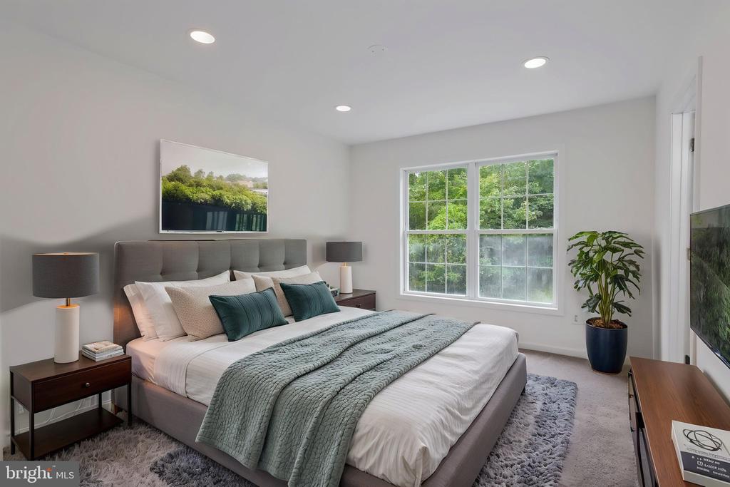 Staged bedroom with a gray upholstered bed, teal accents, and a wide window overlooking trees.
