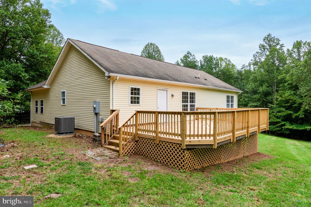 Rear of a beige single-story home with a large raised wooden deck, surrounded by mature trees.