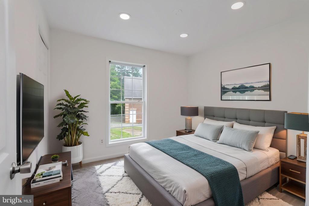 Staged bedroom with a gray upholstered bed, teal throw, large window, and wall-mounted television.