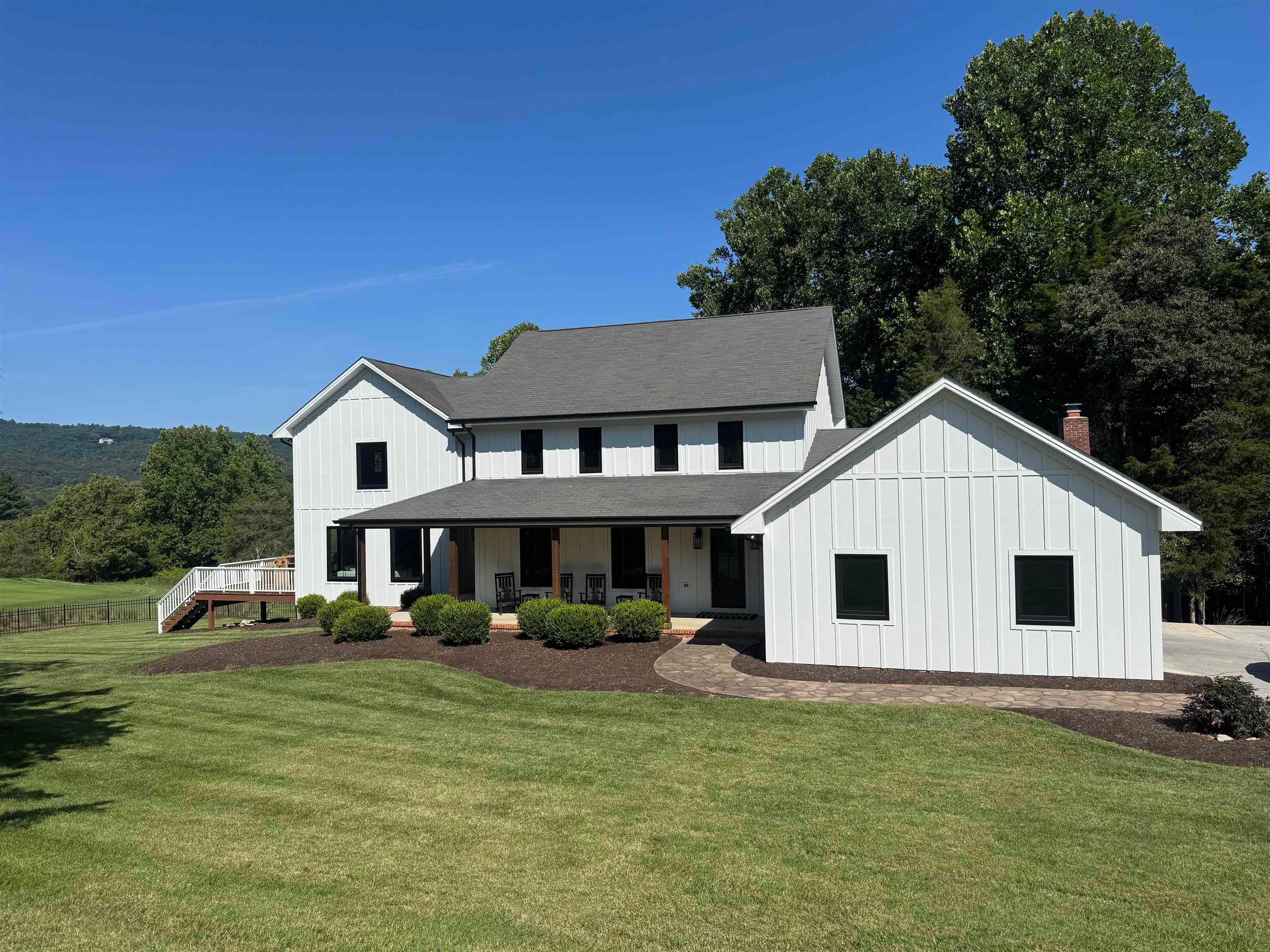 White farmhouse with a covered porch and broad lawn, backed by mature trees and wooded hills.