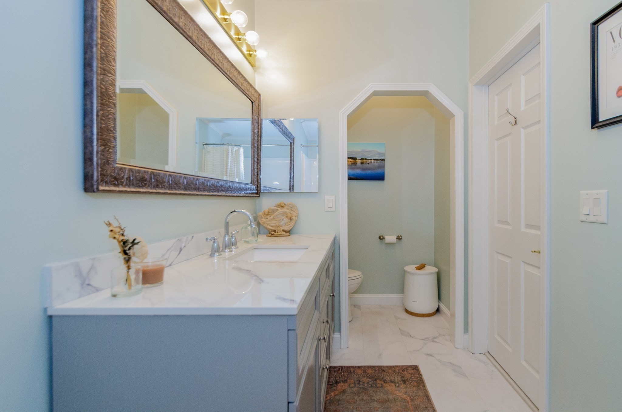 Bathroom with a pale blue vanity and large framed mirror beside a toilet in an arched alcove.