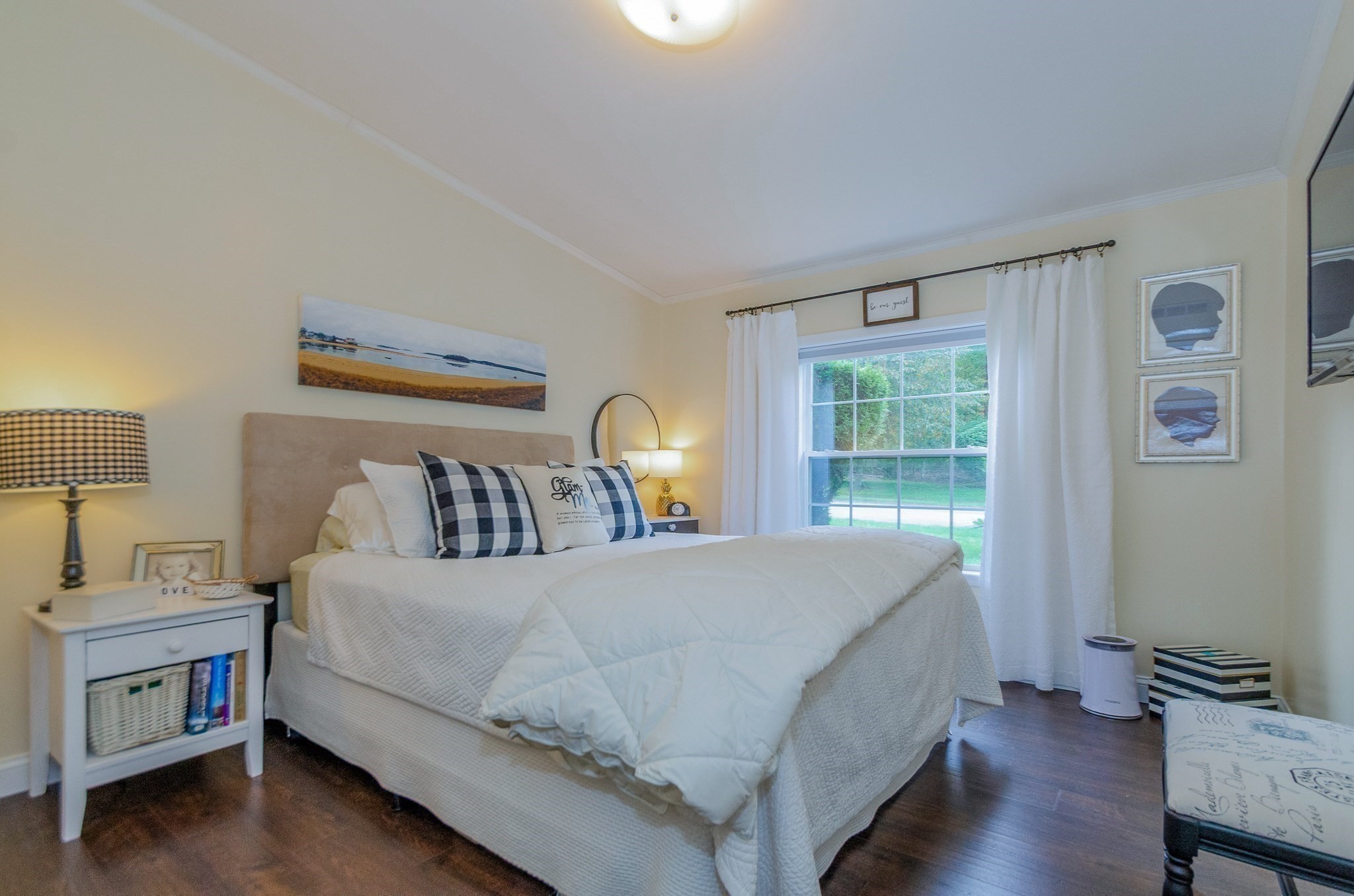 Furnished bedroom with a white bed, plaid pillows, beige headboard, and window overlooking greenery.