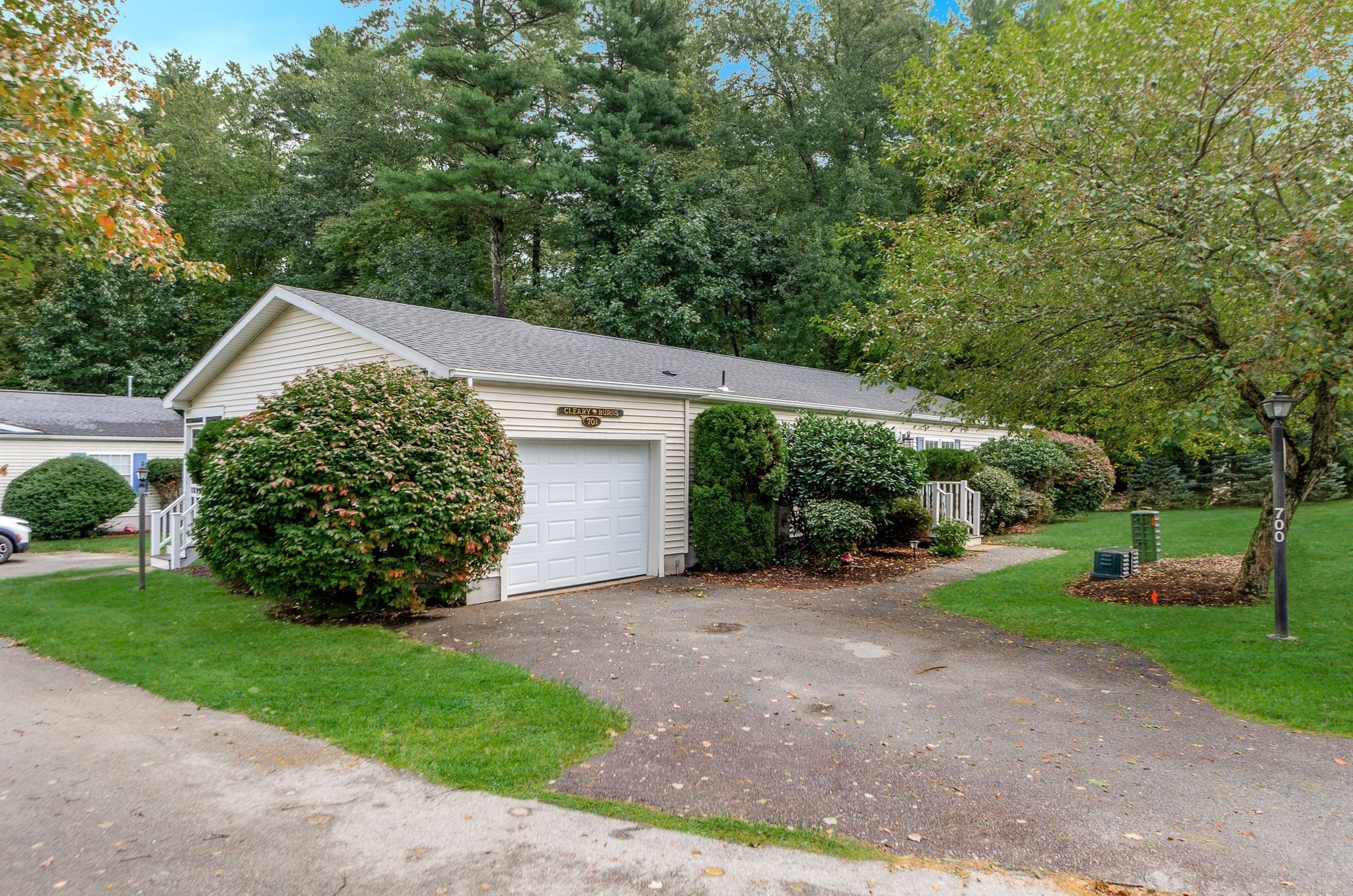 Beige home with a white garage door, landscaped shrubs, and a paved driveway beneath mature trees.