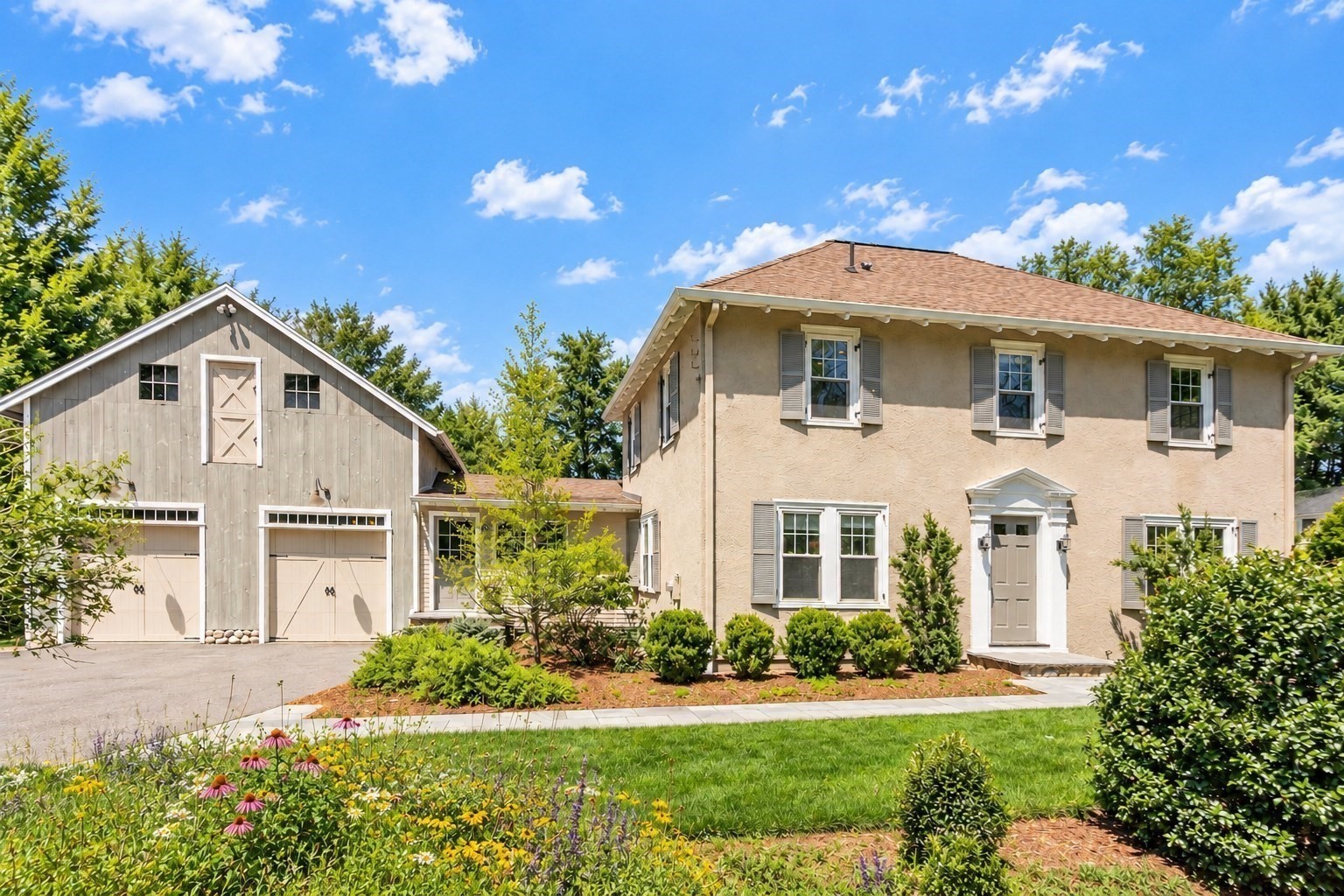 Two-story beige house beside a gray barn-style garage, with flower beds and a landscaped lawn.