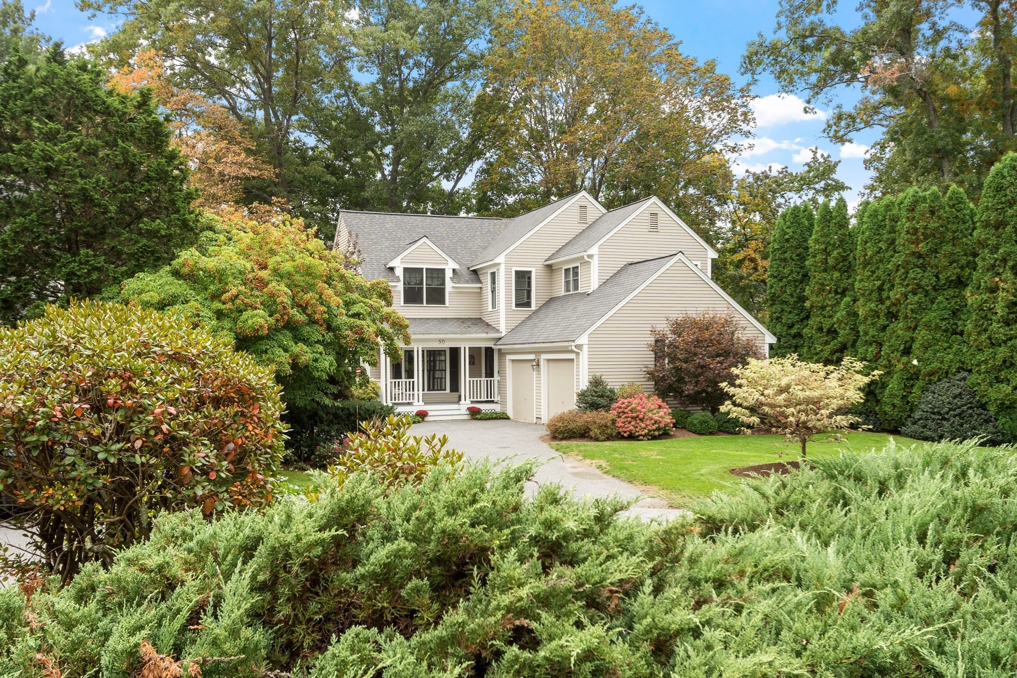 Beige two-story house with a covered porch, attached garage, and mature autumn landscaping.