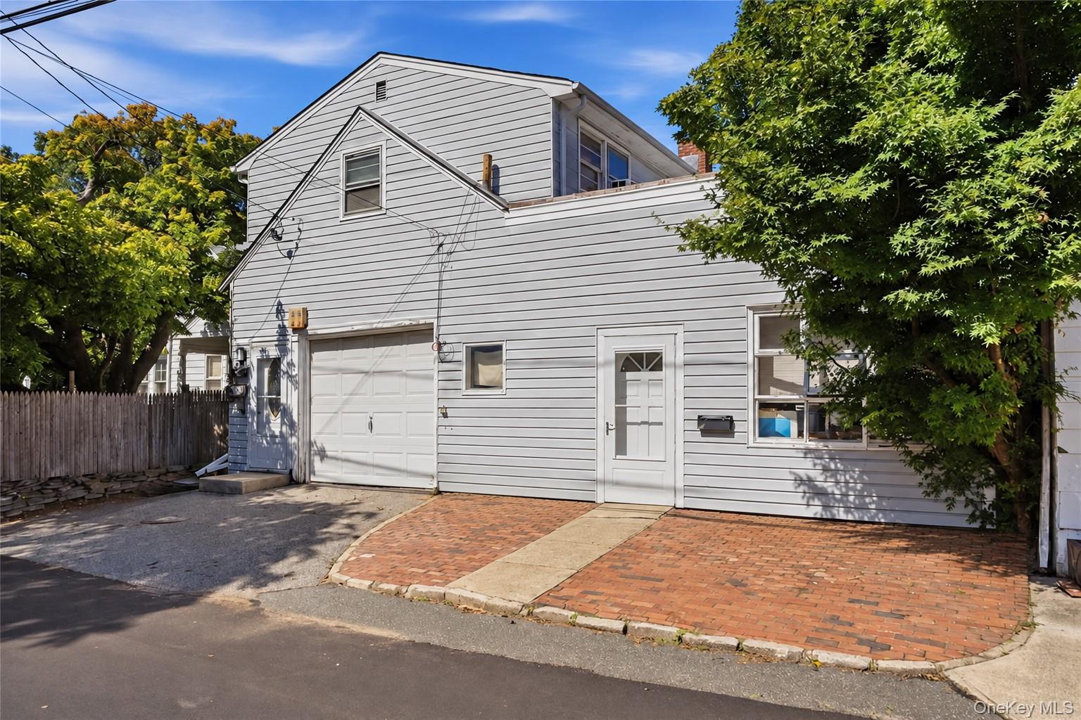 White-sided two-story home with a garage and brick driveway beneath mature trees.