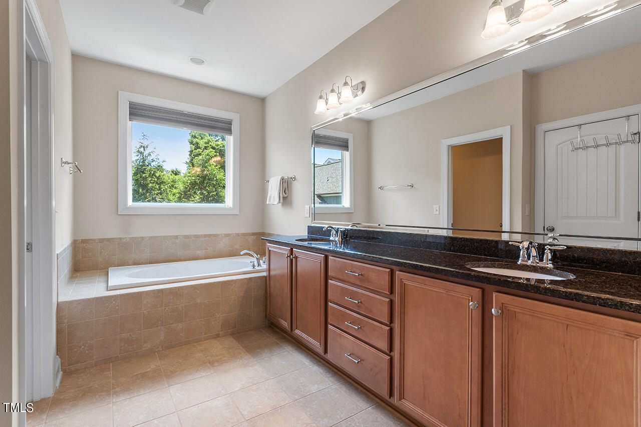 Bathroom with a tiled bathtub beneath a window and a long double vanity with dark countertops.