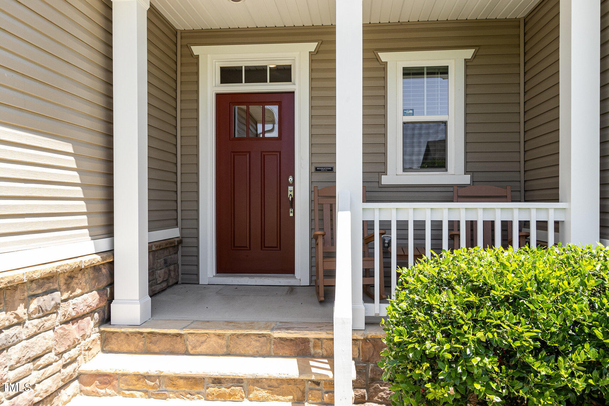 Home’s front porch with a dark red door, stone steps, wooden chairs, and a green hedge.