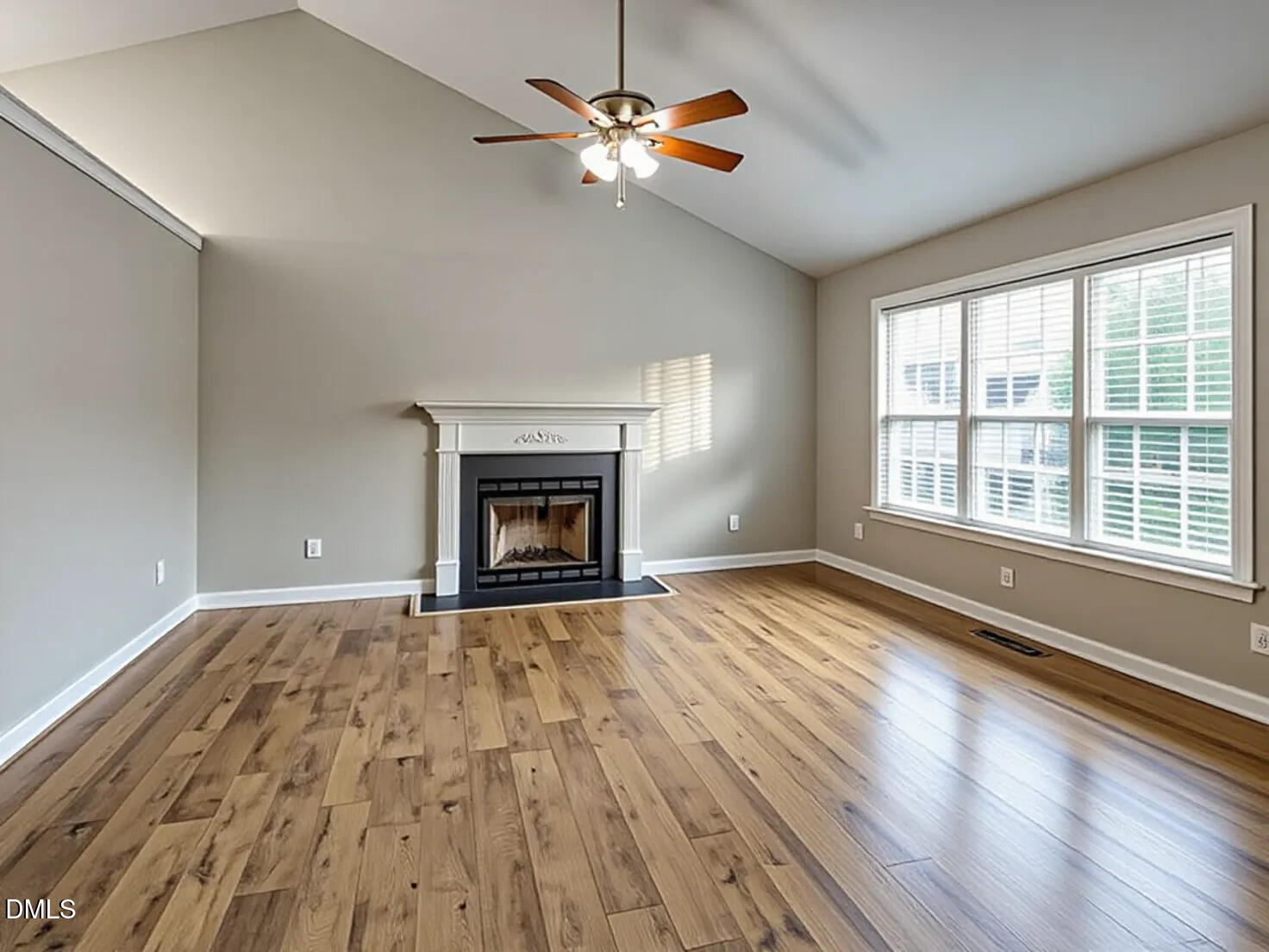 Vacant living room with a fireplace, vaulted ceiling, large windows, and wood plank flooring.
