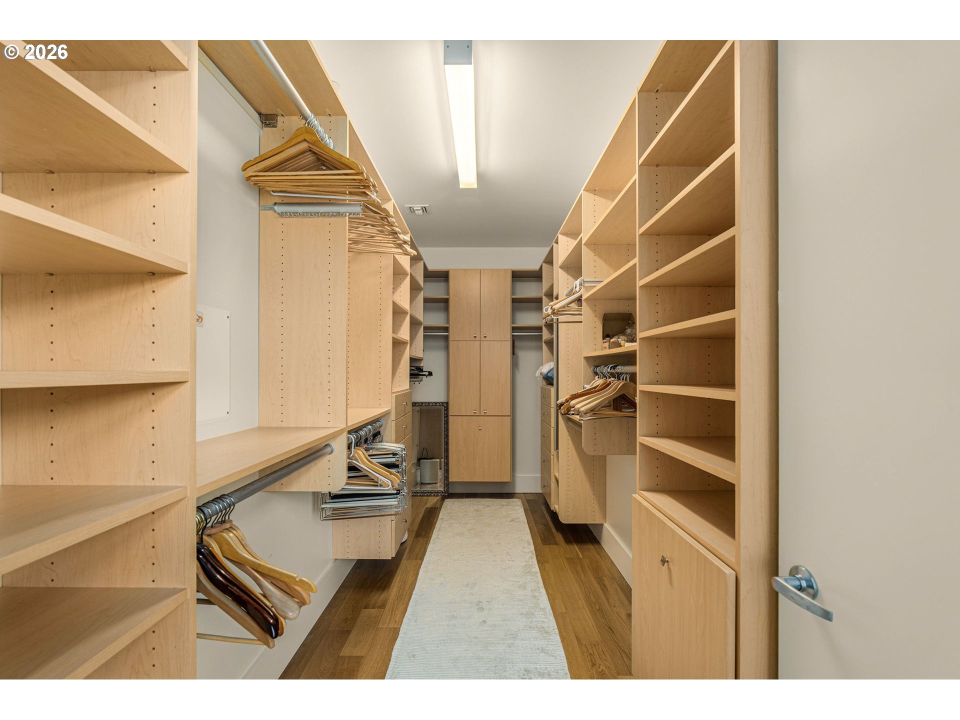 Walk-in closet with light wood built-ins, hanging rods, cabinets, and a pale runner rug.