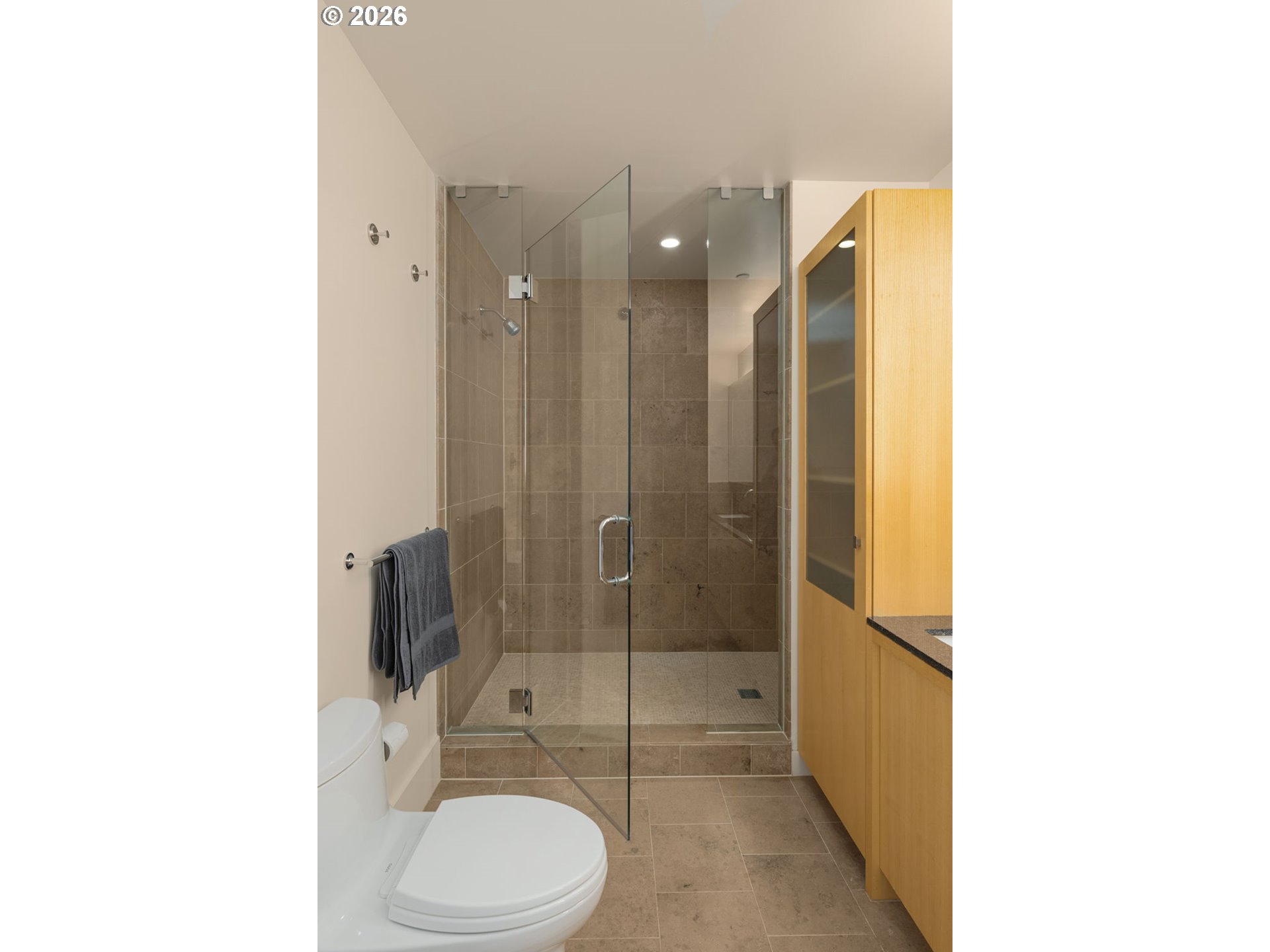 Condo bathroom with a glass walk-in shower, beige tile, and light wood cabinetry.