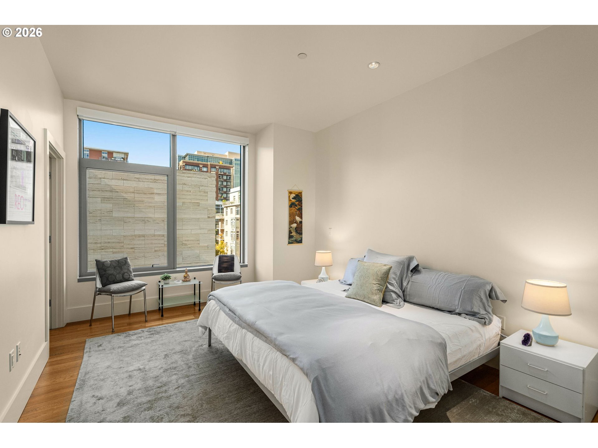 Furnished bedroom with a large window overlooking nearby city buildings.