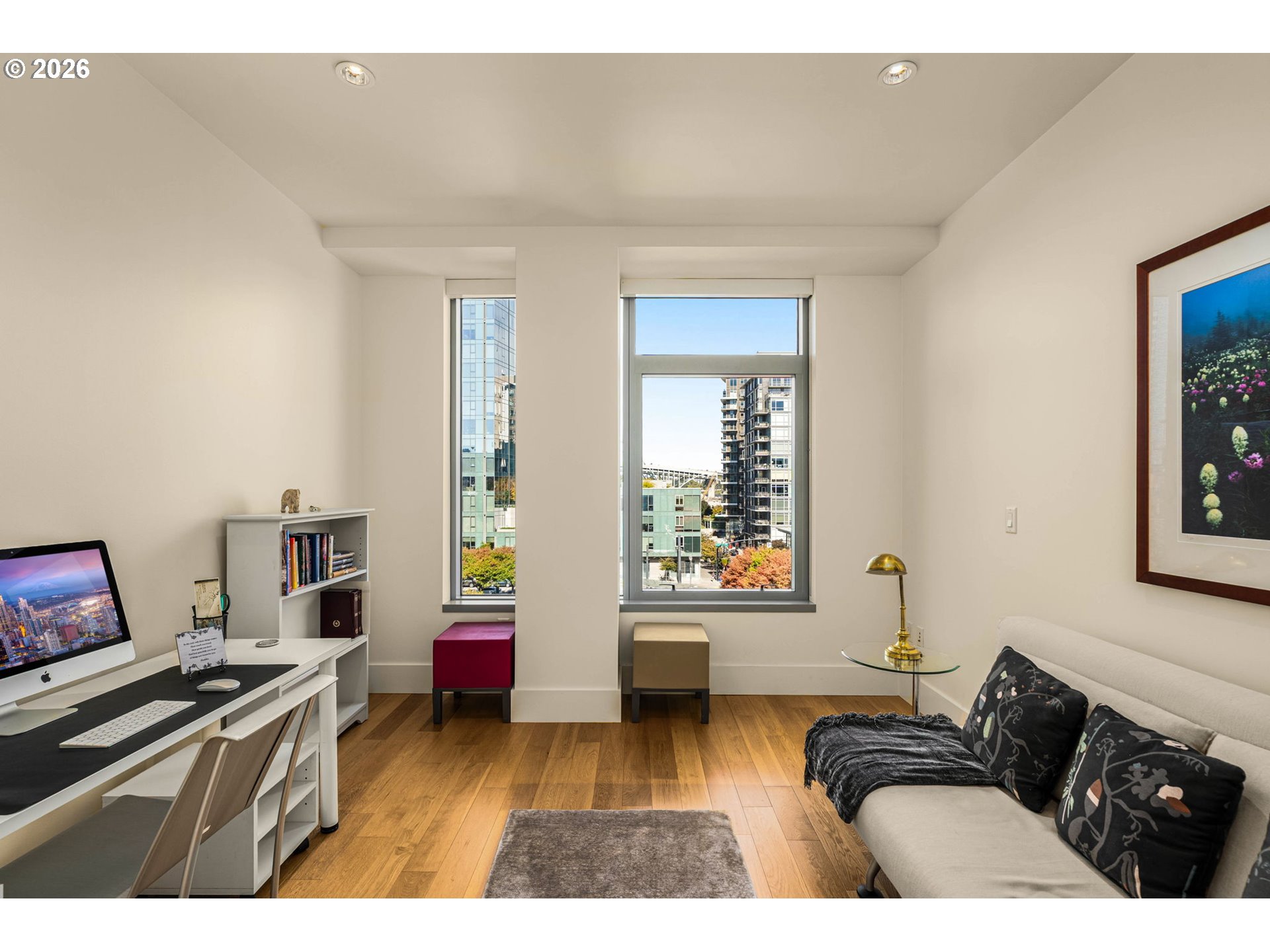 Furnished condo office with a desk, sofa, and tall windows overlooking nearby buildings.