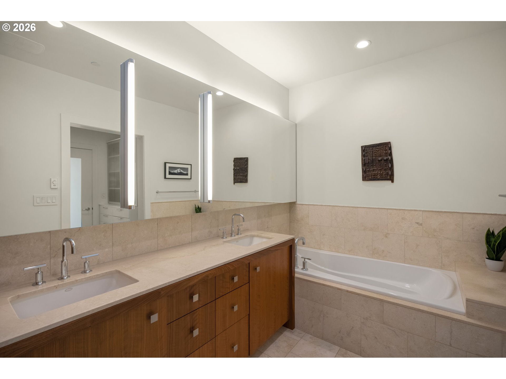 Bathroom with a long double vanity, wood cabinetry, mirrored wall, and built-in tub.