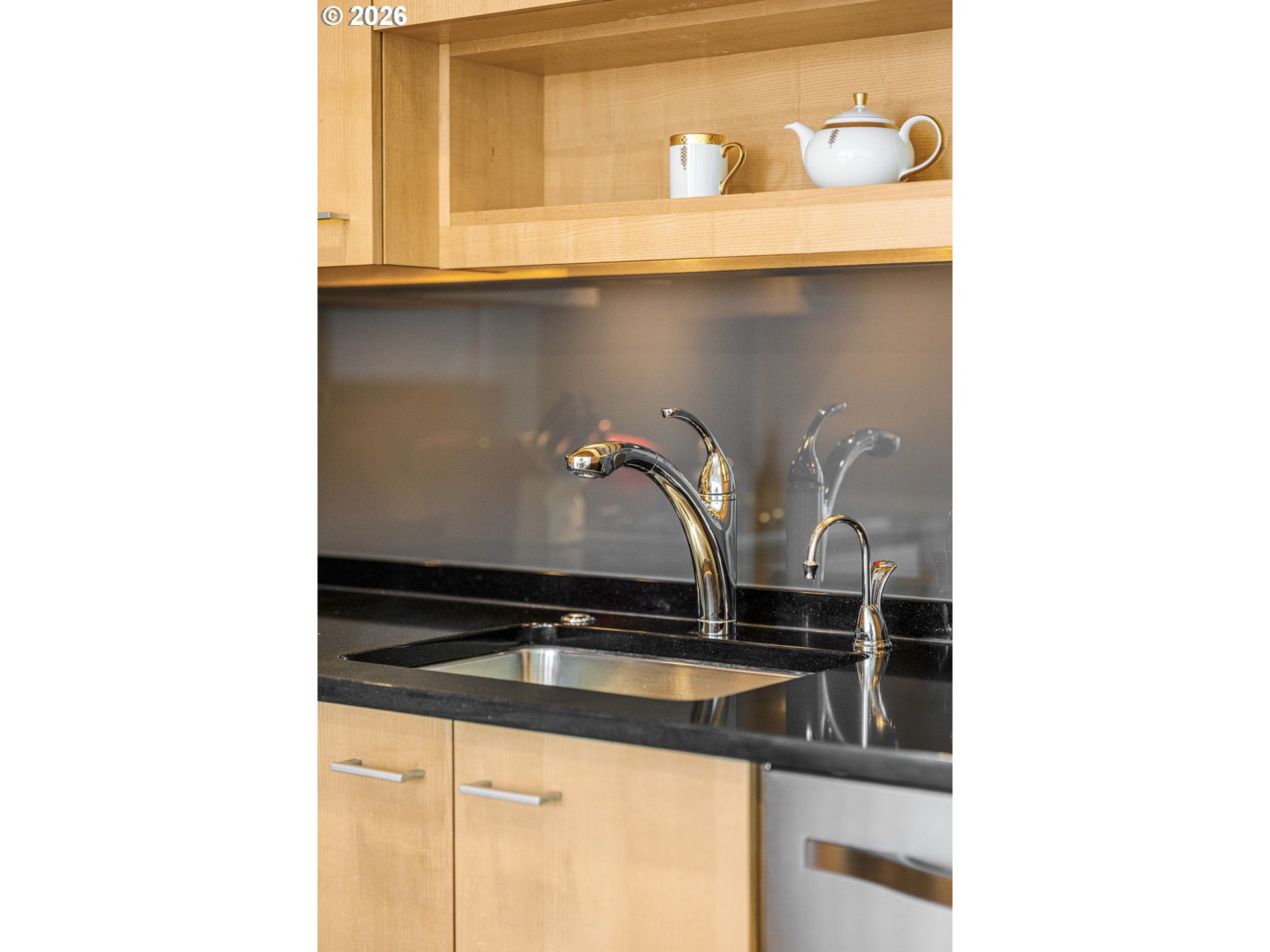 Kitchen sink with polished chrome faucets, black countertop, light wood cabinets, and teapot on an open shelf.
