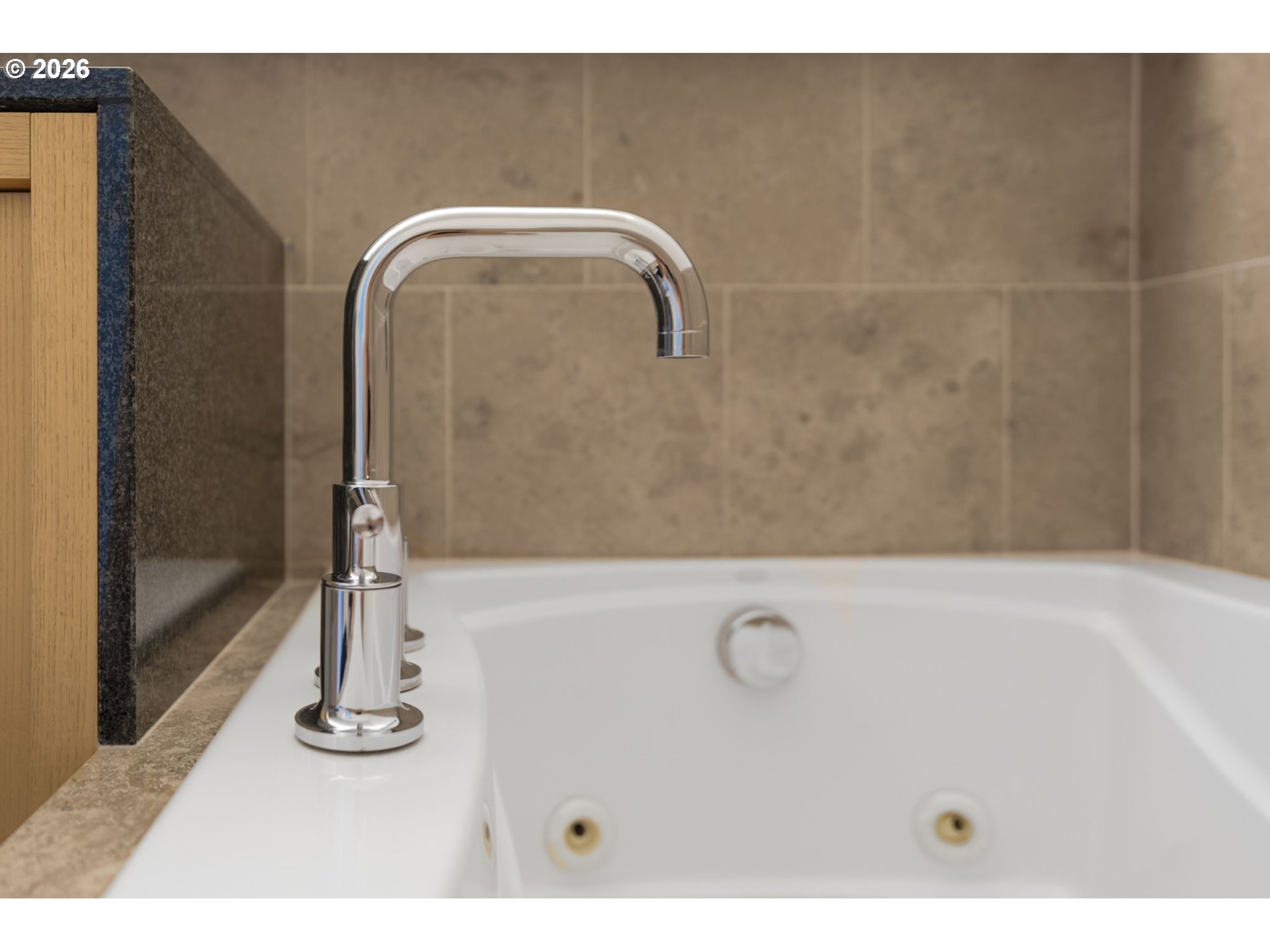 Jetted bathtub with a tall chrome faucet beside a wood vanity and beige tile.
