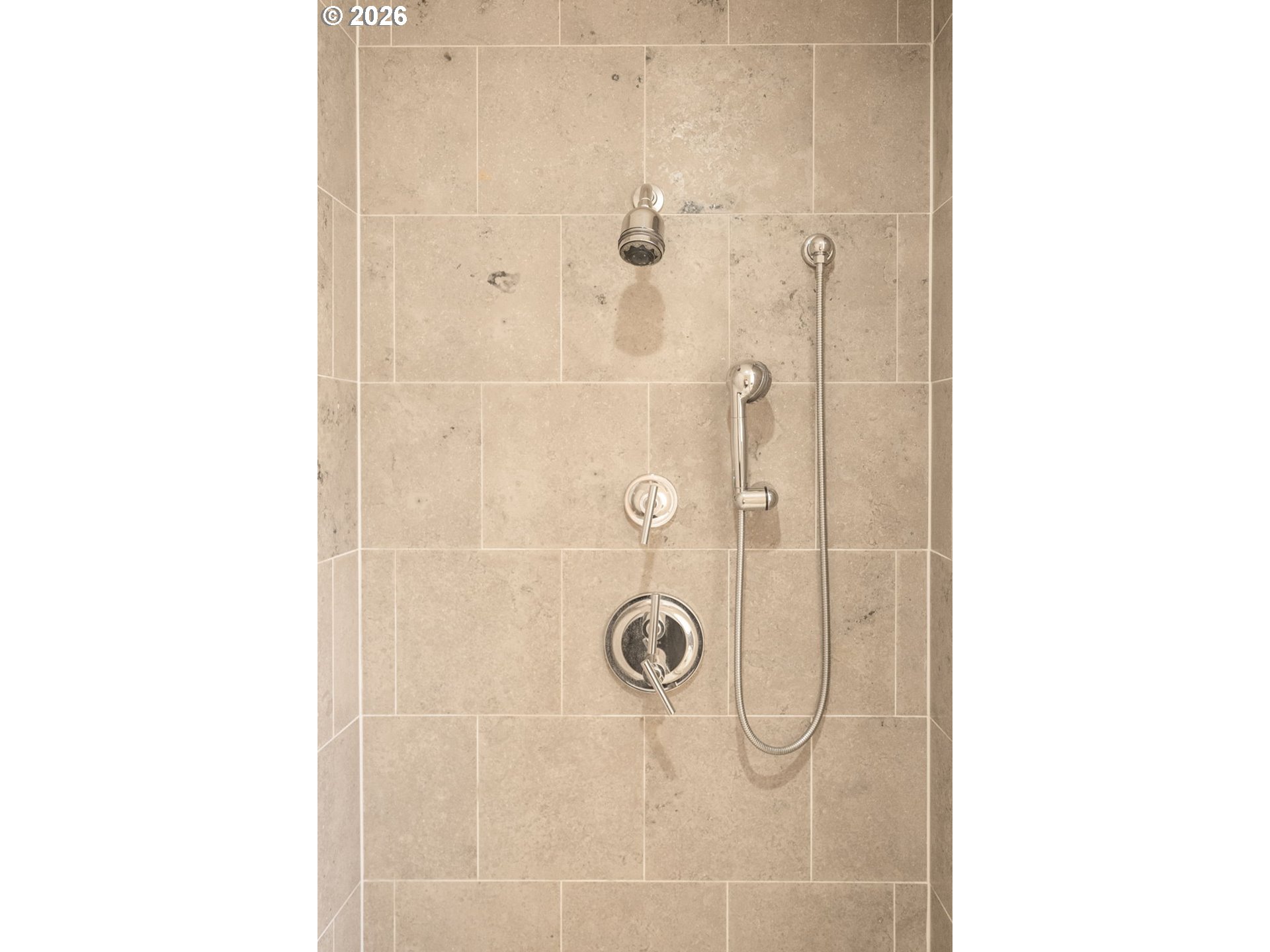 Bathroom shower with beige tile, polished chrome fixtures, and a handheld shower.
