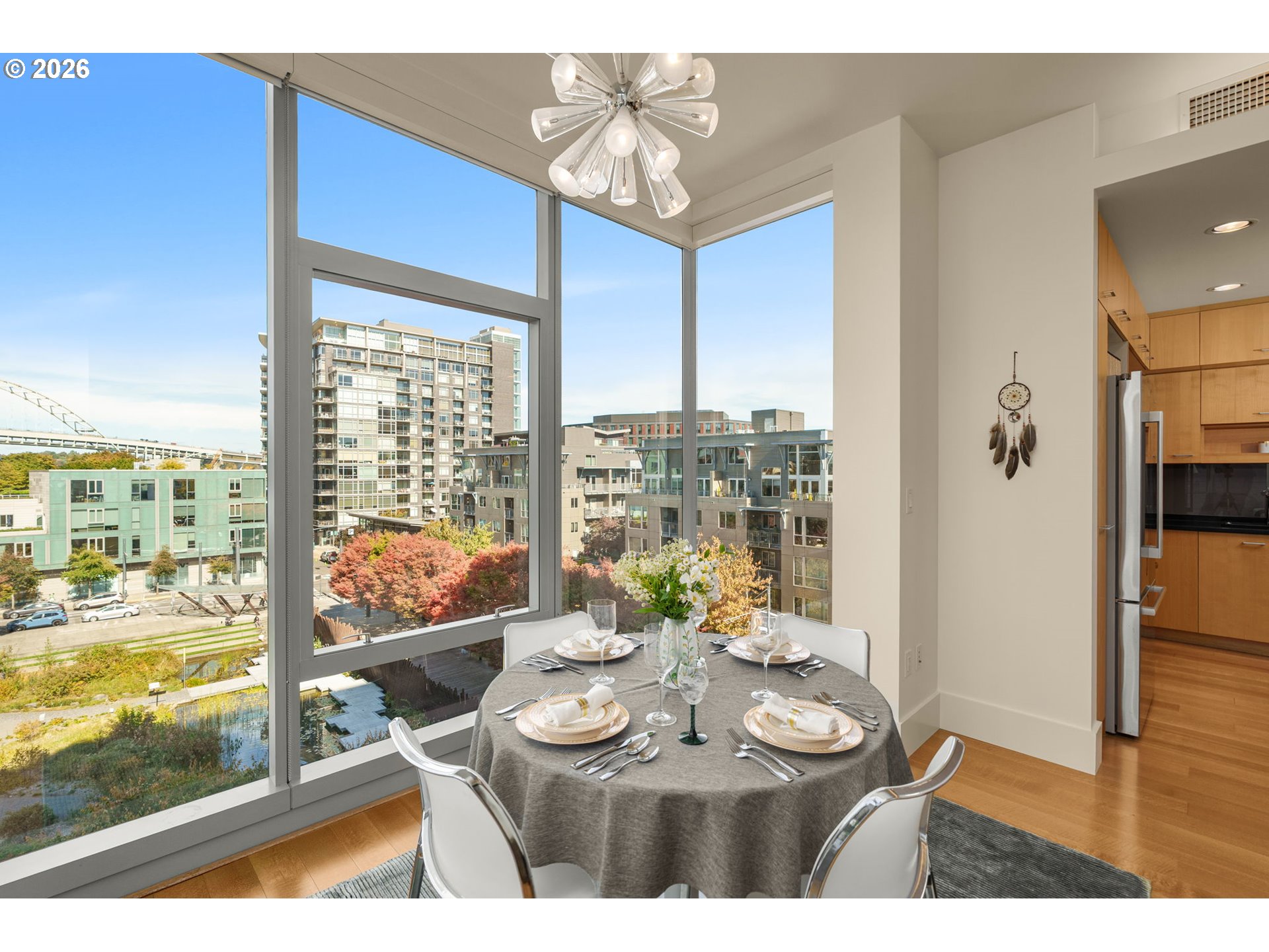 Condo dining area with a set table beside floor-to-ceiling windows overlooking city buildings and autumn trees.