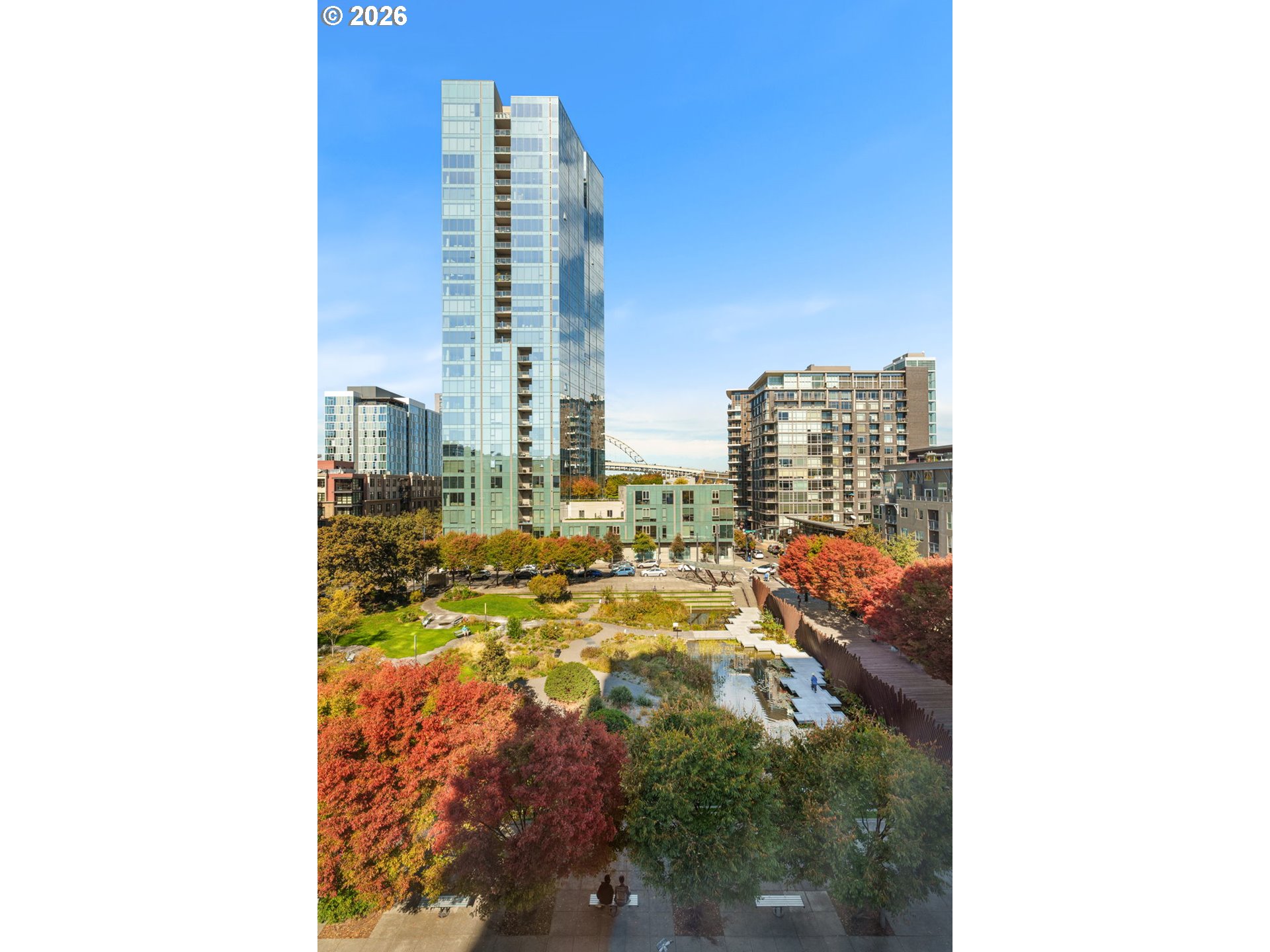 Elevated view over an autumn courtyard toward modern apartment towers, with a water feature and bridge in the distance.