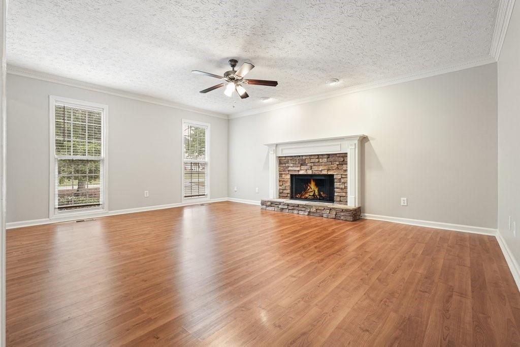 Empty living room with hardwood floors, a stone fireplace, two tall windows, and a ceiling fan.