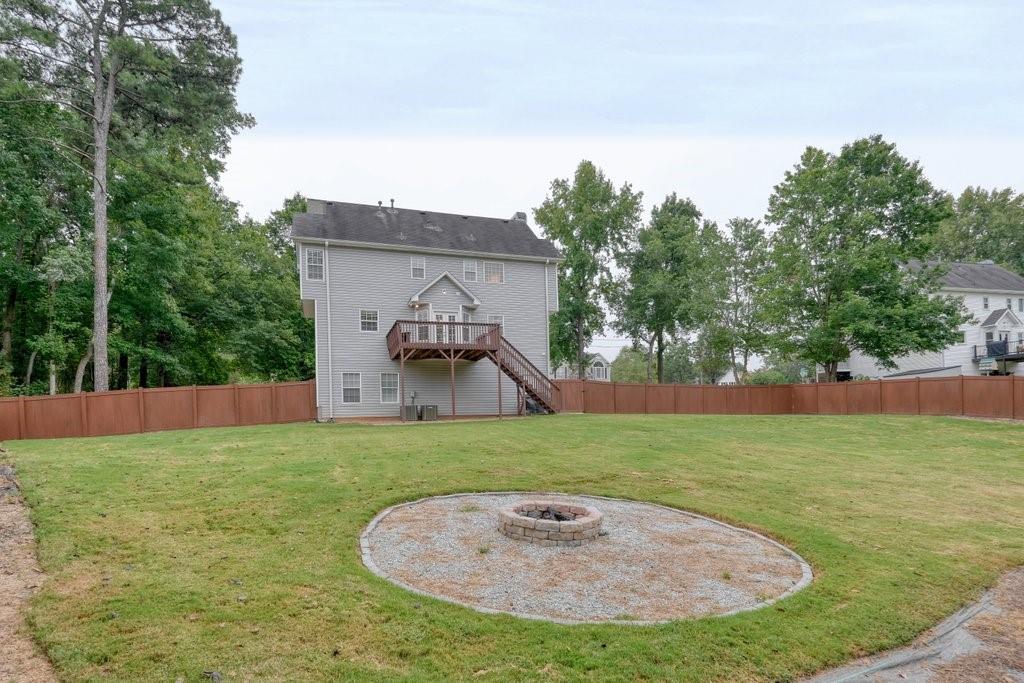 Two-story gray house with a raised deck, fenced lawn, and circular fire-pit area.