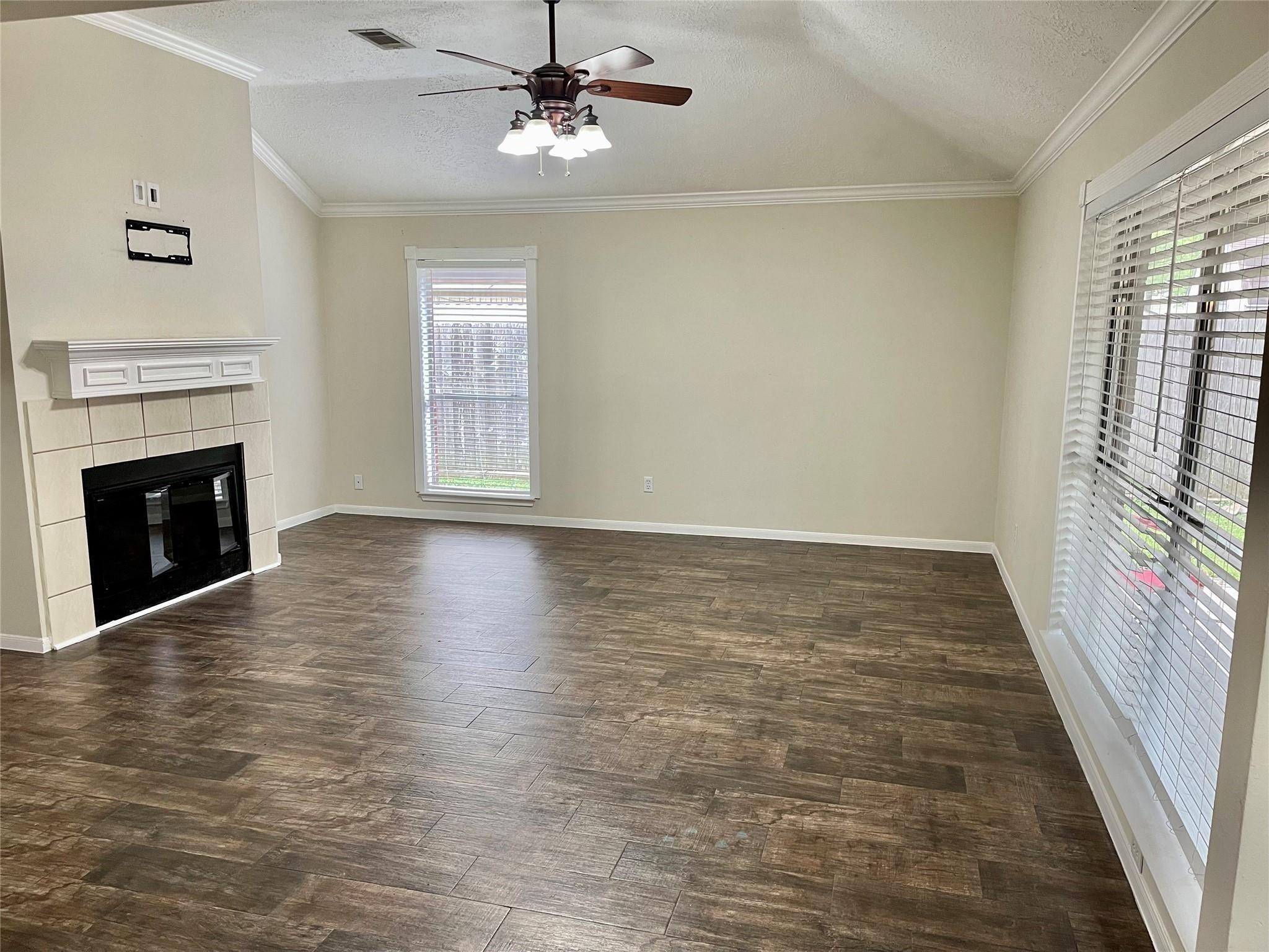 Empty living room with a tiled fireplace, dark wood-look floors, and large windows with blinds.