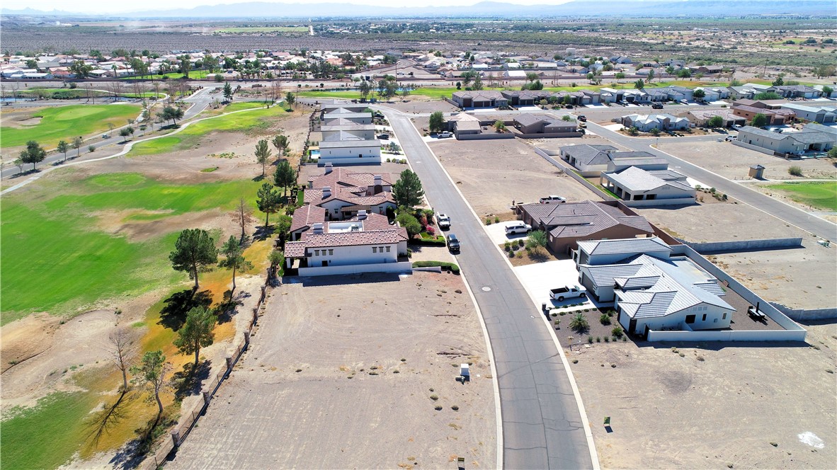 Aerial view of a vacant desert lot beside homes and a green golf course, with mountains beyond.