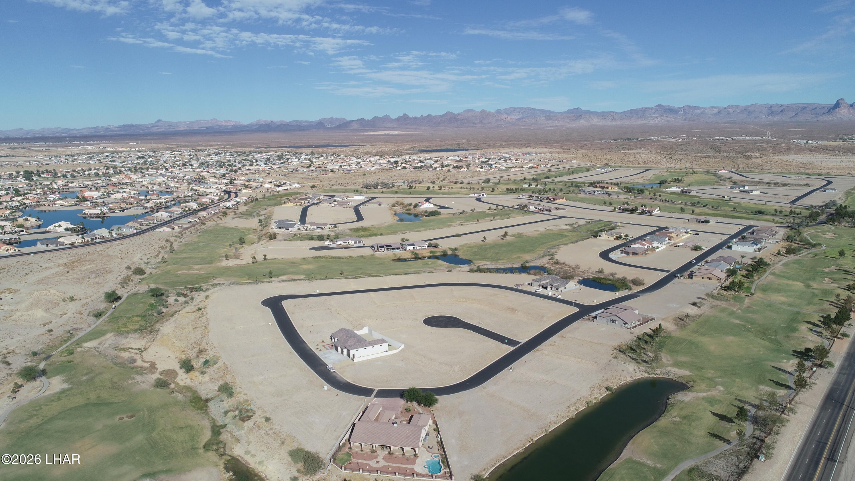 Aerial view of vacant lots and homes beside green golf-course areas and ponds, with desert mountains in the distance.