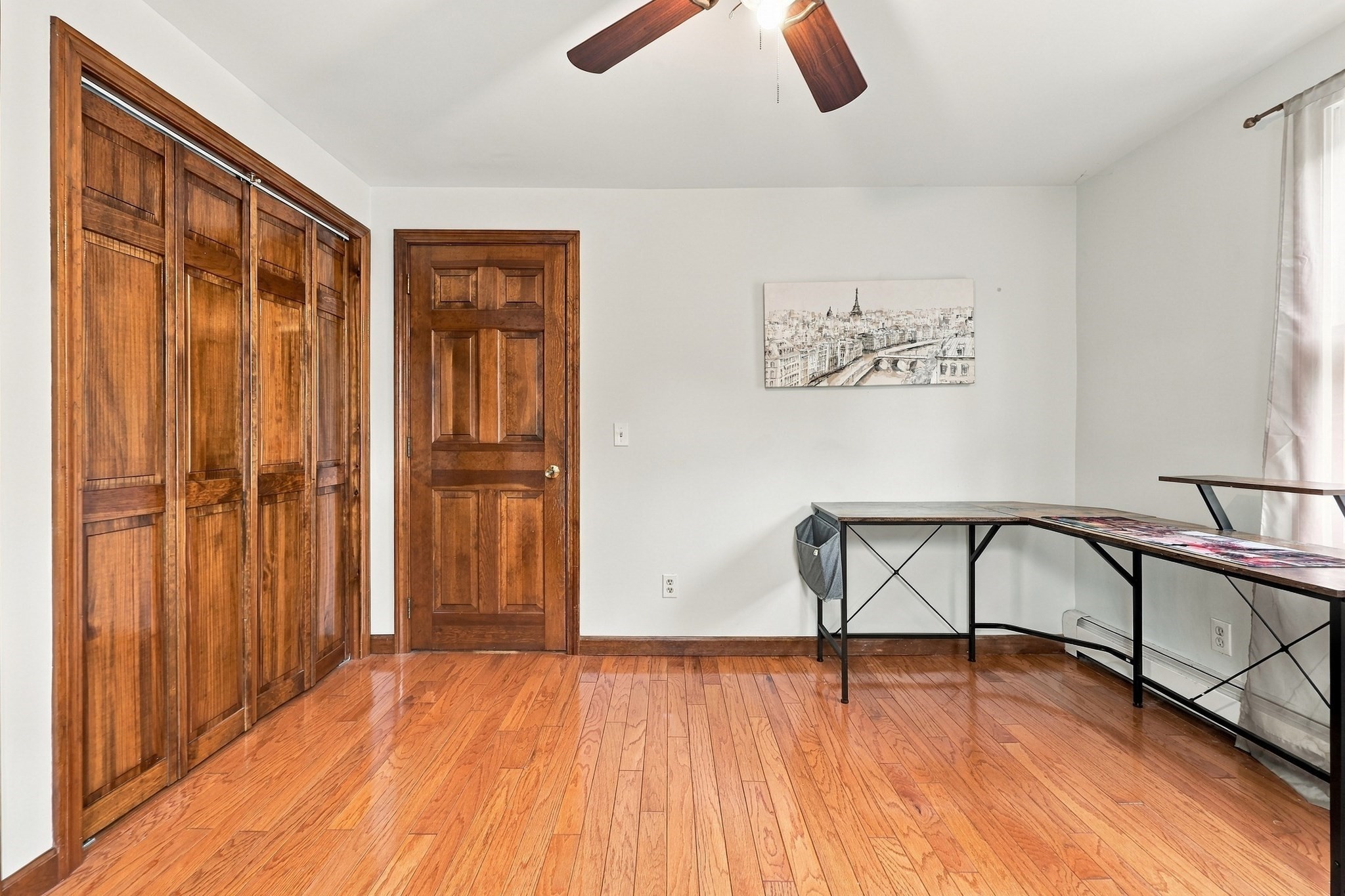 Room with hardwood floors, wooden closet doors, and a desk beneath a framed cityscape print.