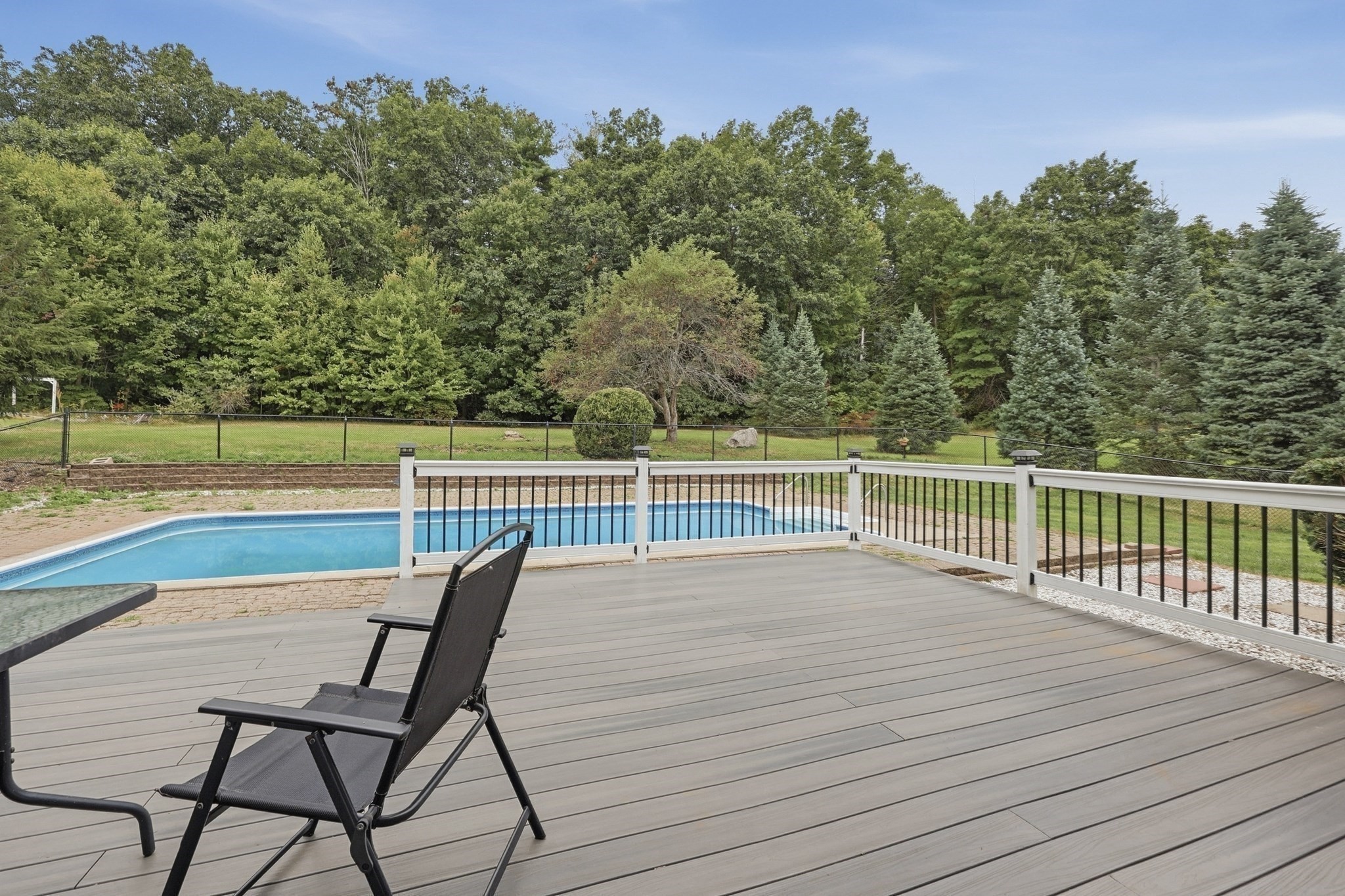 Gray deck and black patio chair overlook a swimming pool, fenced yard, and wooded backdrop.