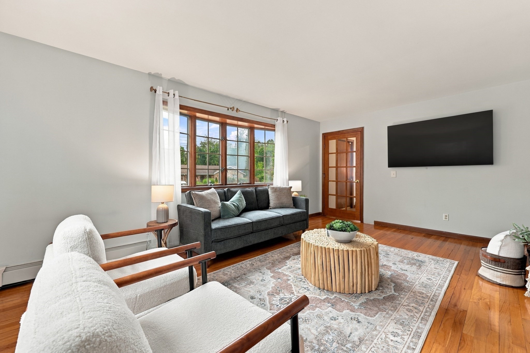 Living room with hardwood floors, a large wood-framed window, gray sofa, and wall-mounted television.