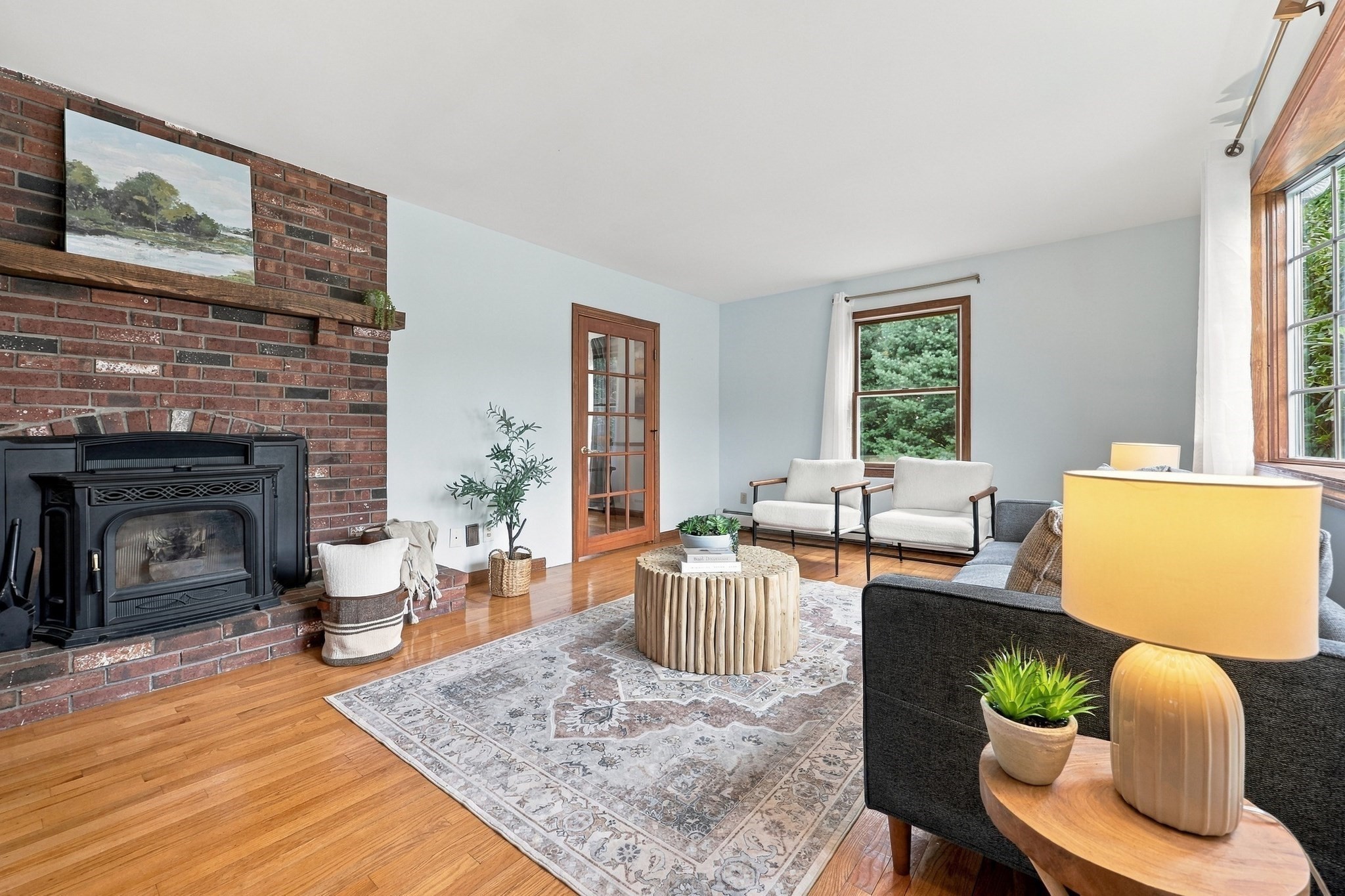 Living room with a brick fireplace, wood stove, hardwood floors, and seating by large windows.