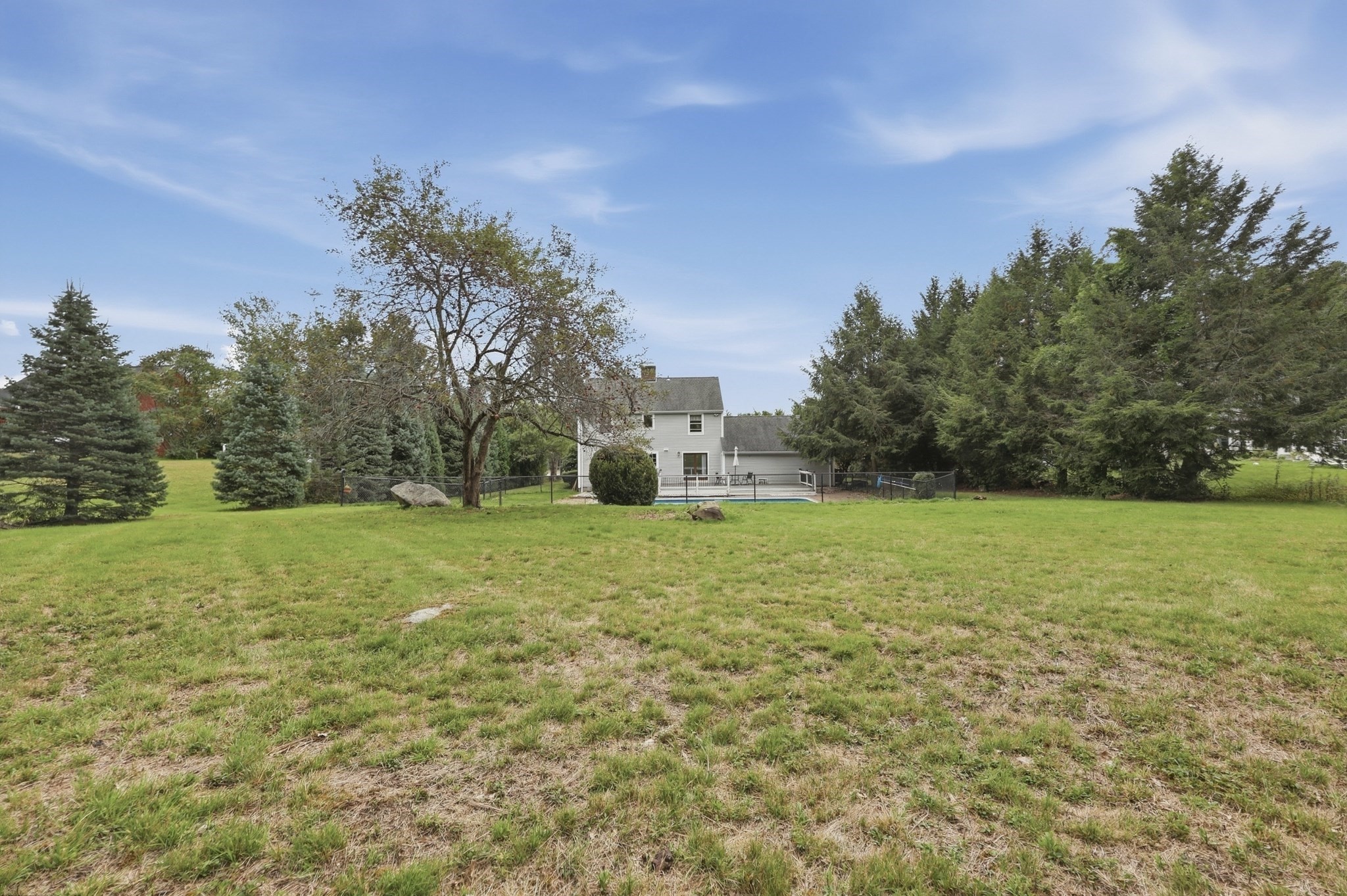 Gray two-story house and fenced pool beyond a large grassy backyard lined with mature trees.