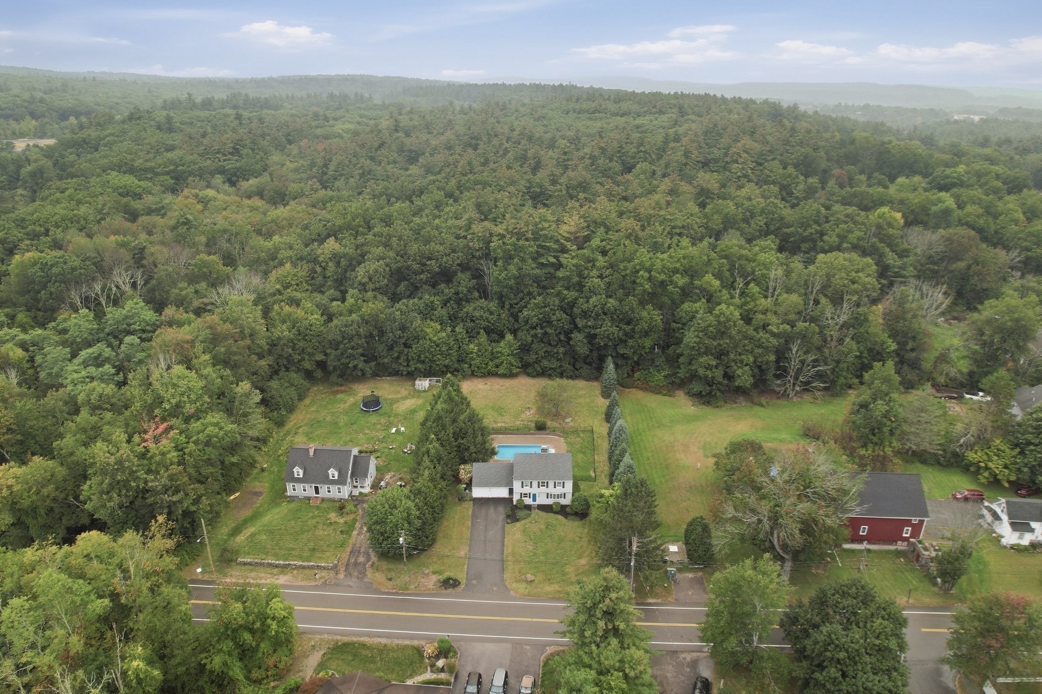 Aerial view of a white house with a swimming pool, long driveway, and woods behind it.