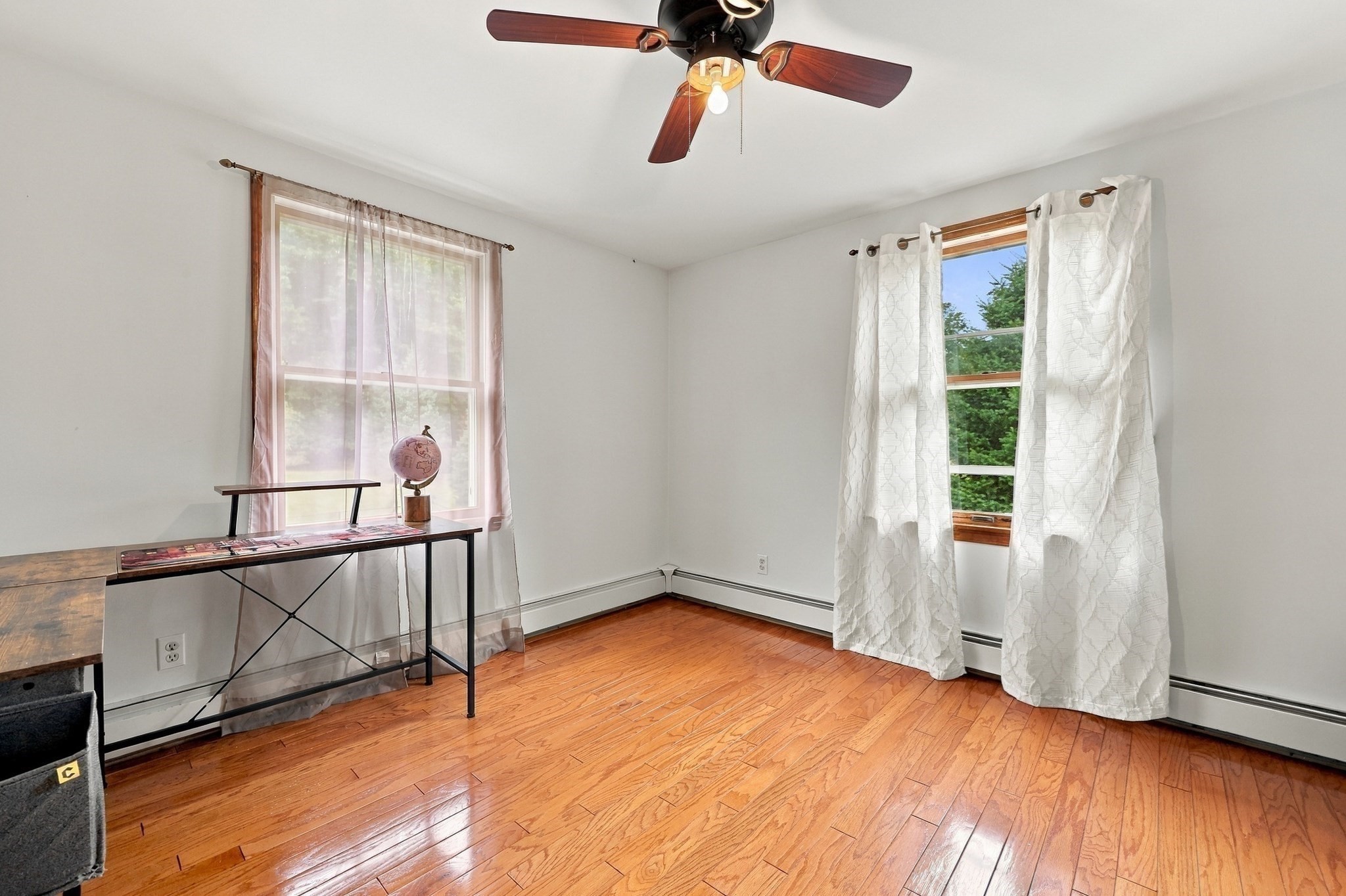 Bright room with polished hardwood floors, two curtained windows, a ceiling fan, and a narrow desk with a globe.