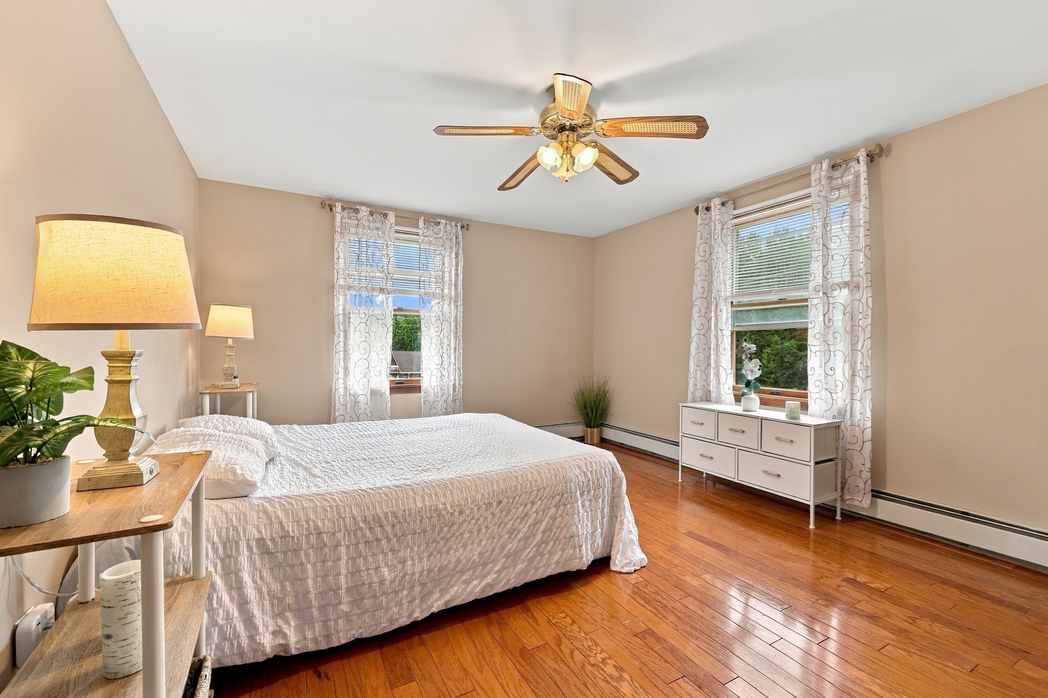 Bedroom with a white bed, polished hardwood floors, two curtained windows, and a ceiling fan.