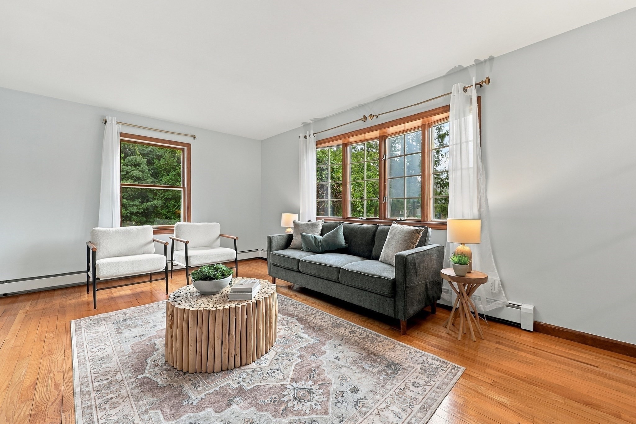 Living room with a dark gray sofa, white armchairs, hardwood floors, and large windows overlooking greenery.
